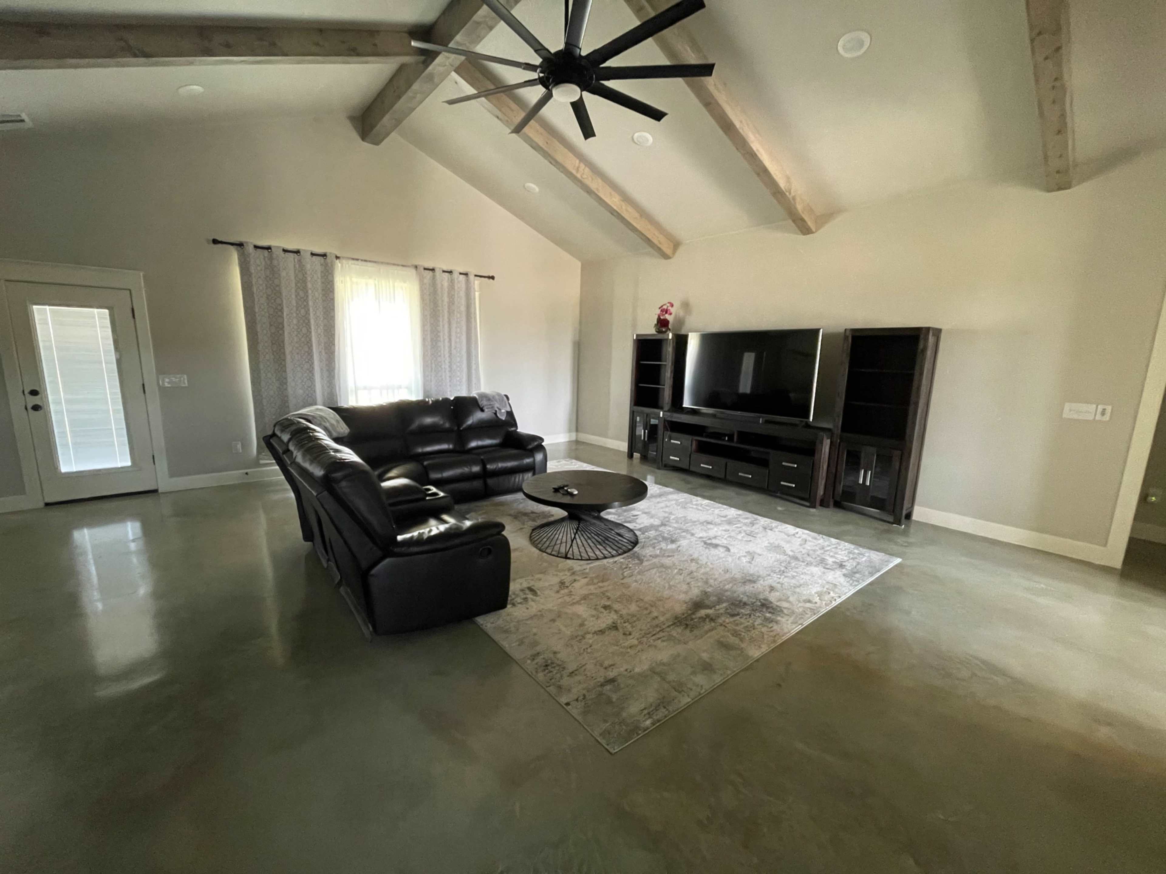 A modern living room with a black leather sofa, a round coffee table on a rug, and a large TV unit against a wall with vaulted ceilings and wooden beams.