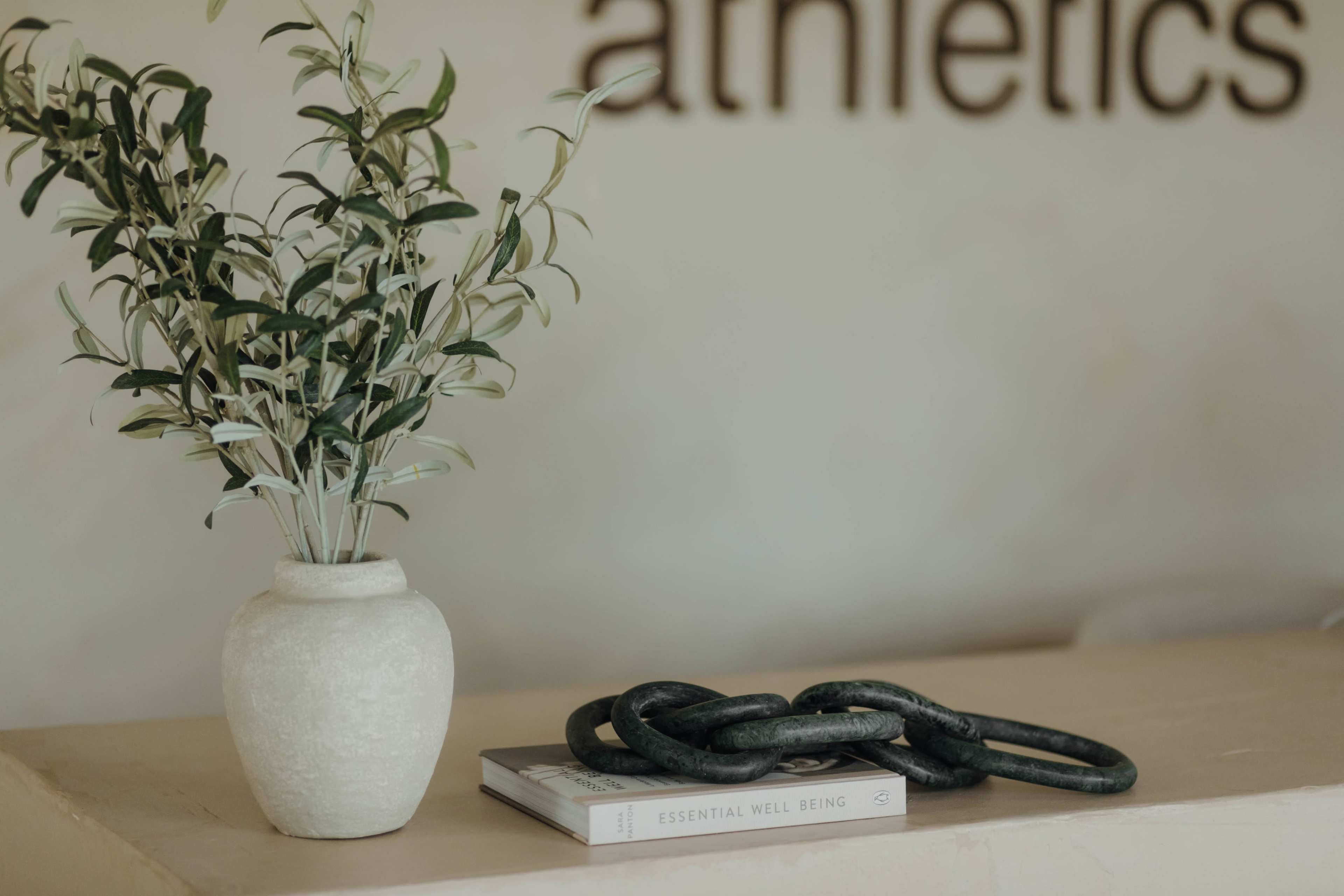A potted plant and a chain sculpture rest on a stack of books on a smooth, light-colored surface in front of a wall displaying the word "athletics."