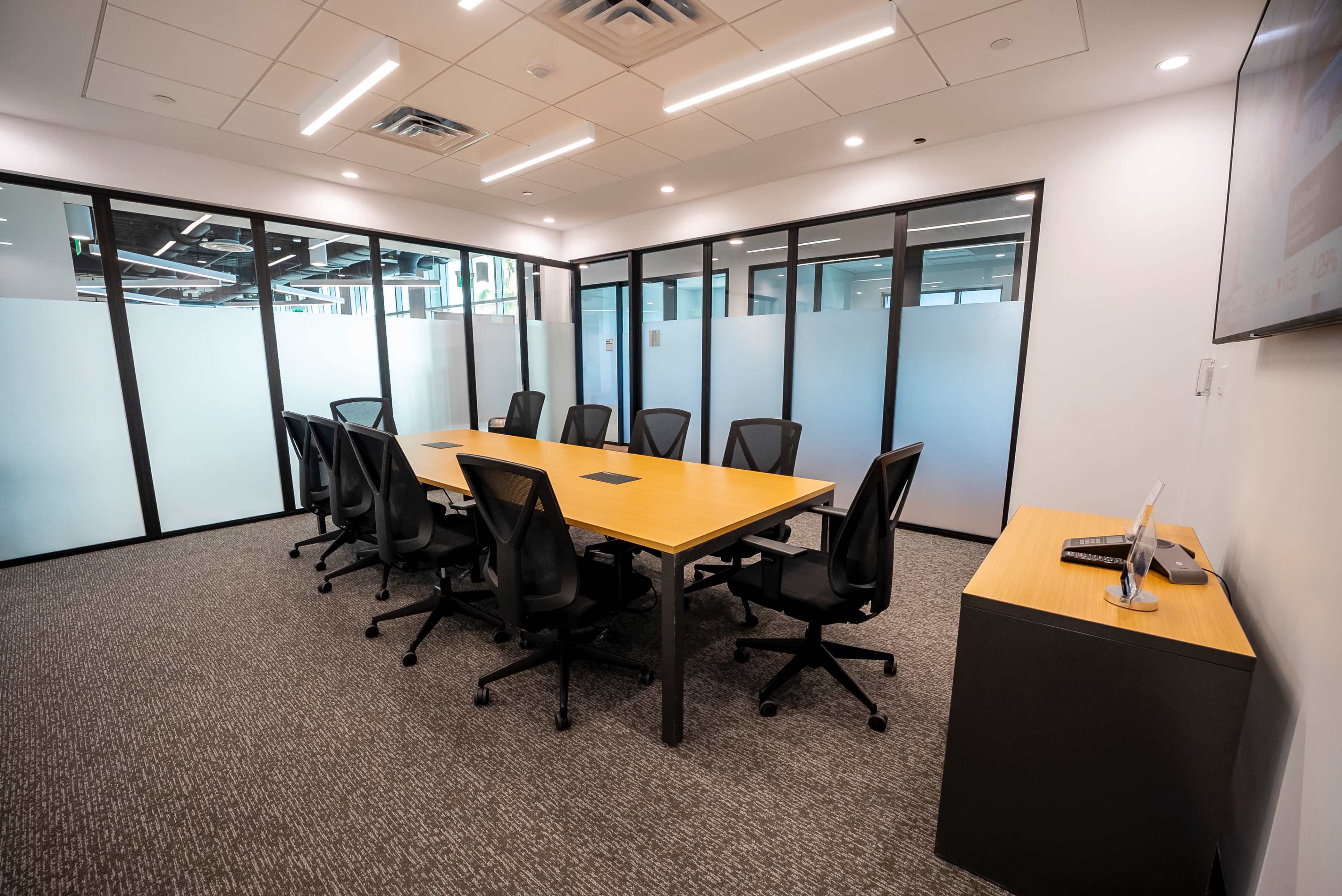 The image shows a modern conference room with a long wooden table surrounded by black office chairs and glass walls.