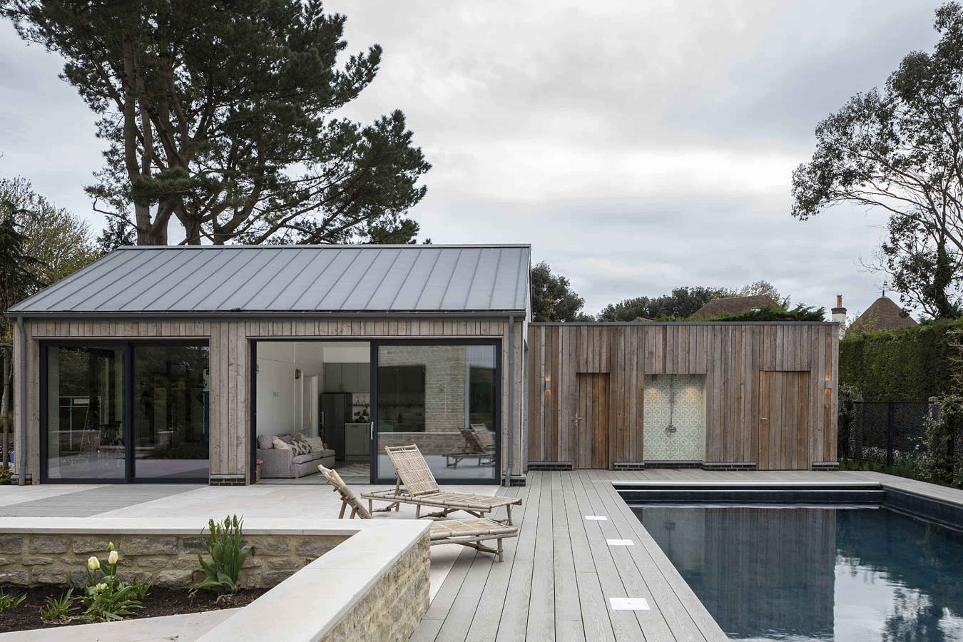 A modern wooden house with a metal roof is situated next to a swimming pool, surrounded by trees and landscaped areas.