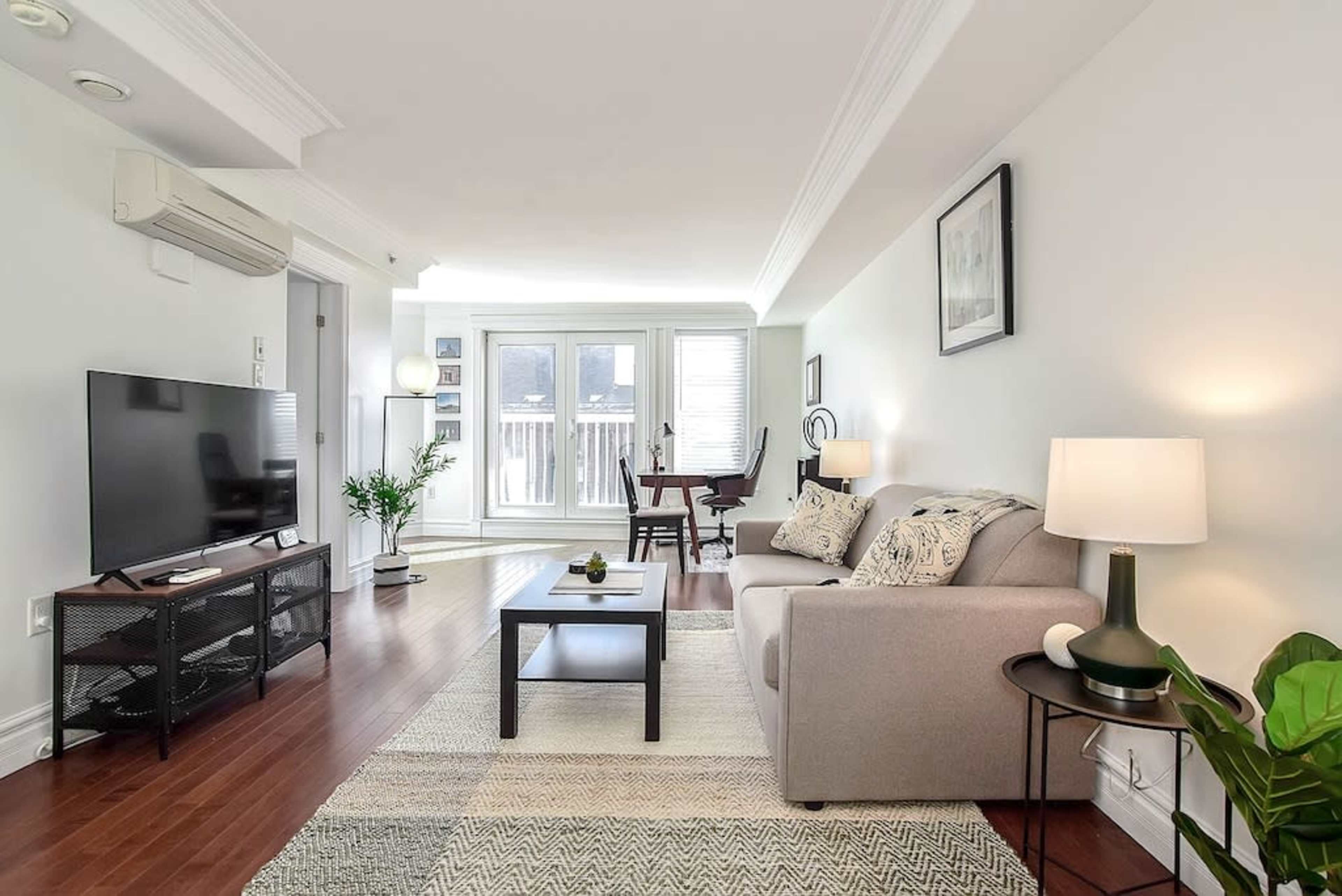 A modern living room featuring a gray sofa, a television on a black stand, and a dining area with a table near large windows.