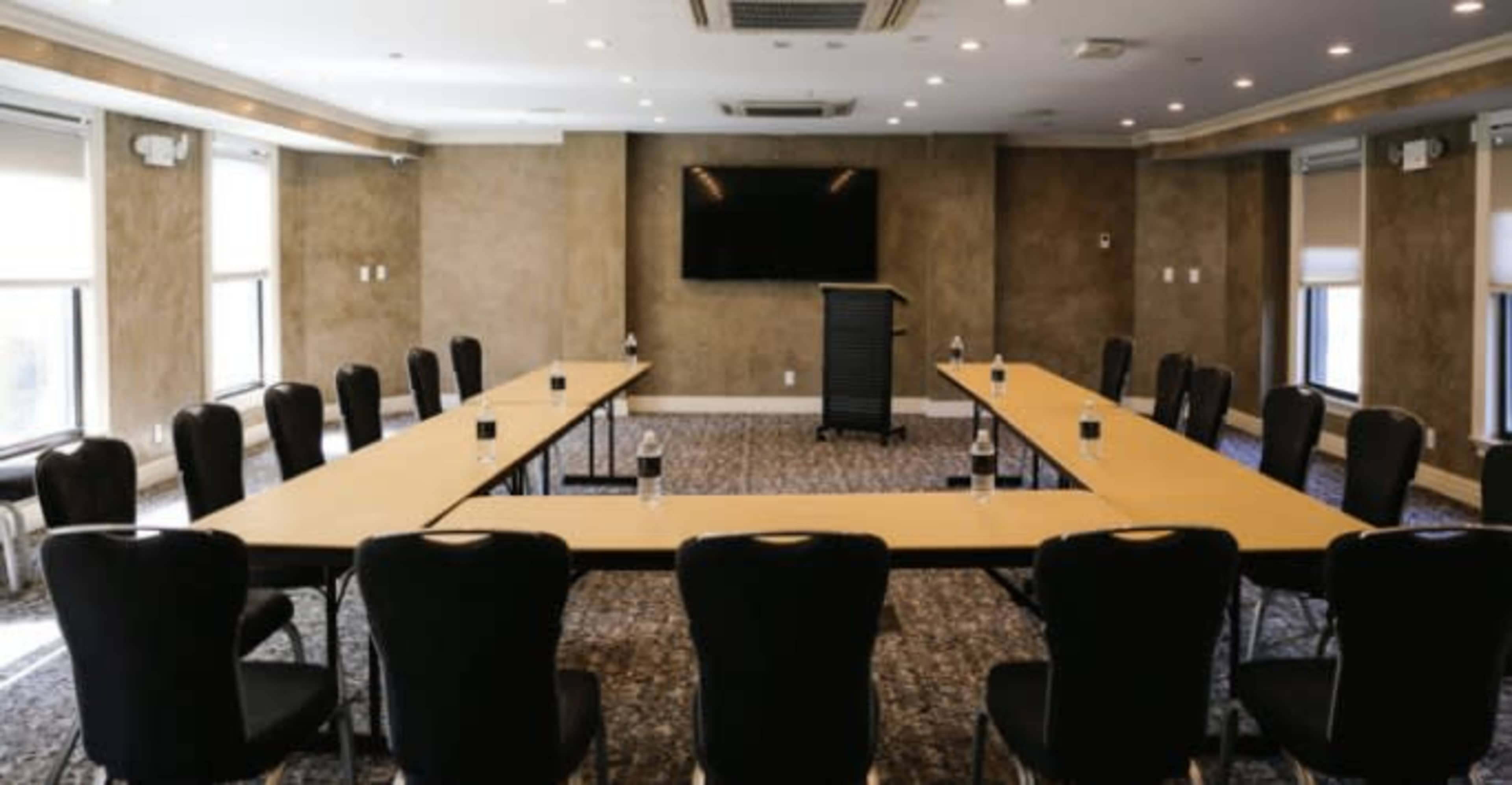 A conference room is arranged with a large oval table surrounded by black chairs, with a podium and a screen at one end.