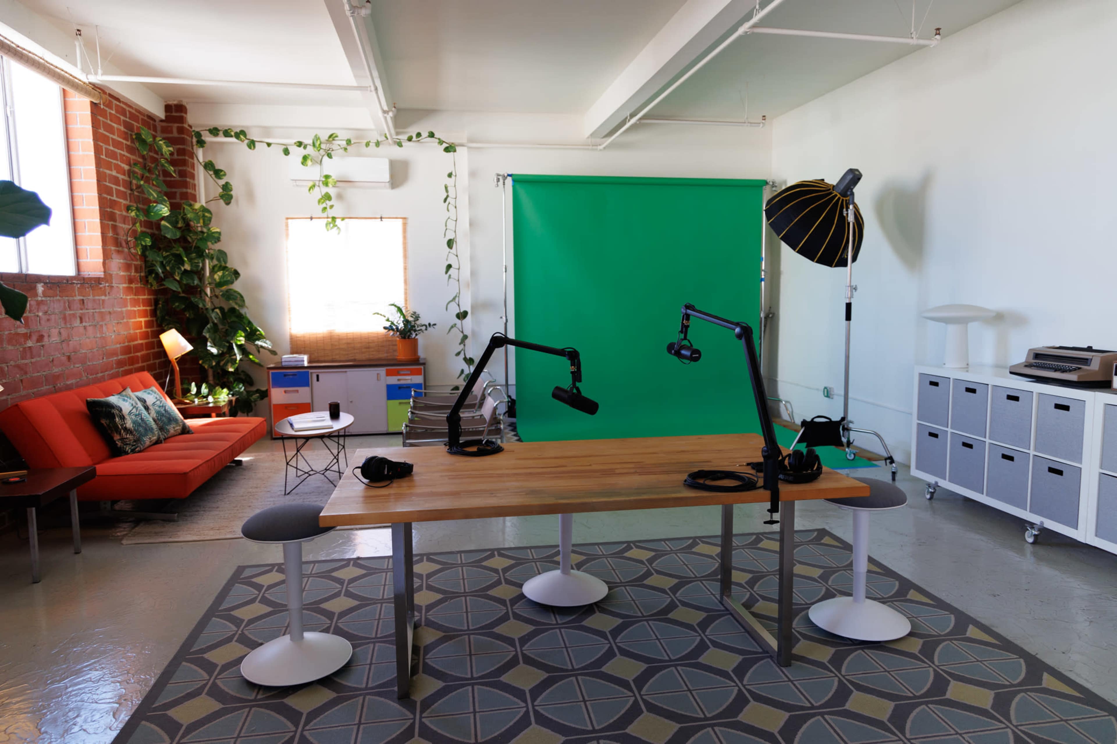 The image shows a modern studio setup featuring a wooden table with two microphone stands, headphones, a green backdrop, and a seating area with a red couch and plants.