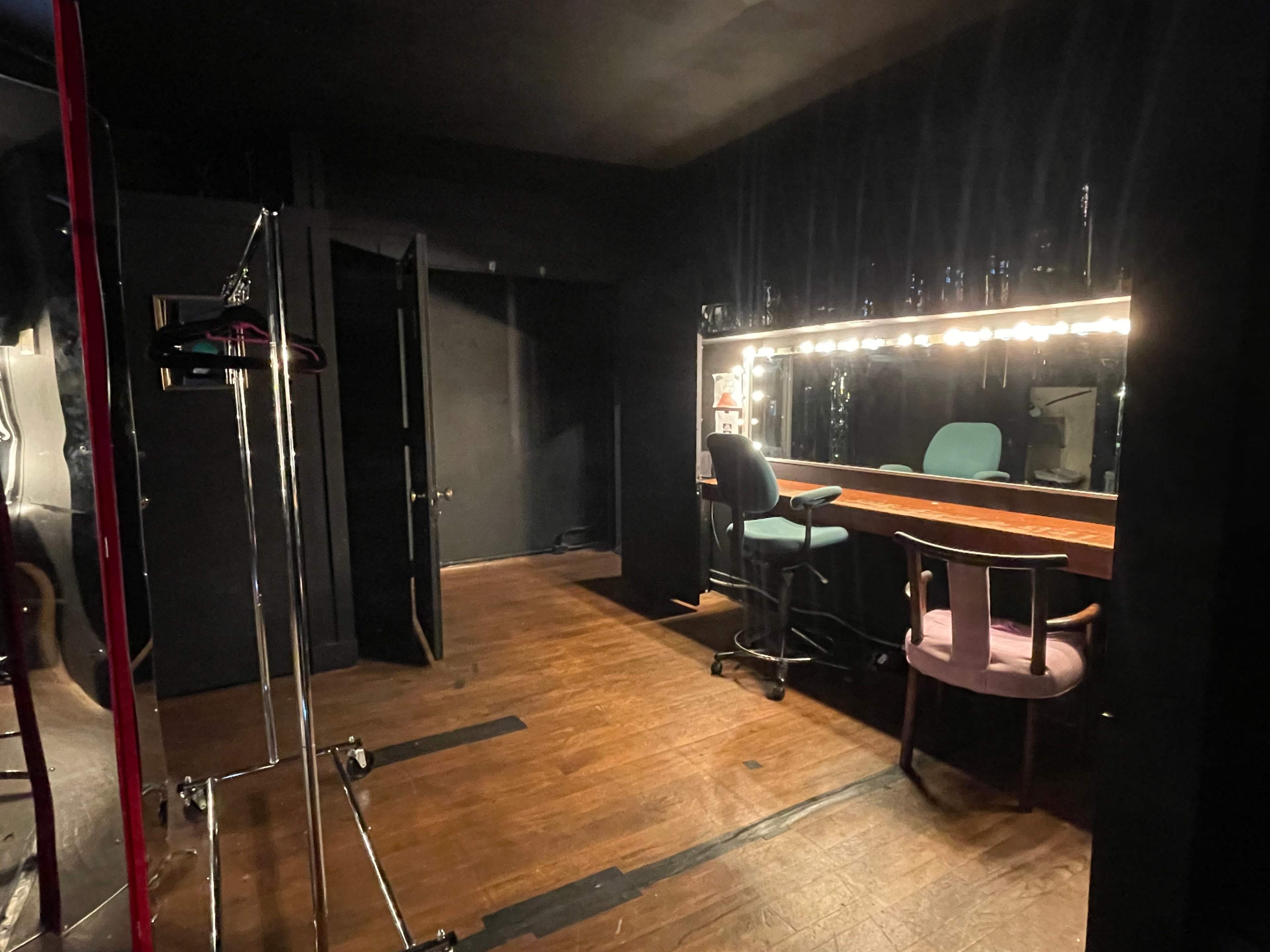 A dimly lit dressing room featuring a mirror with lights, a wooden table, and chairs, along with a clothing rack in one corner.
