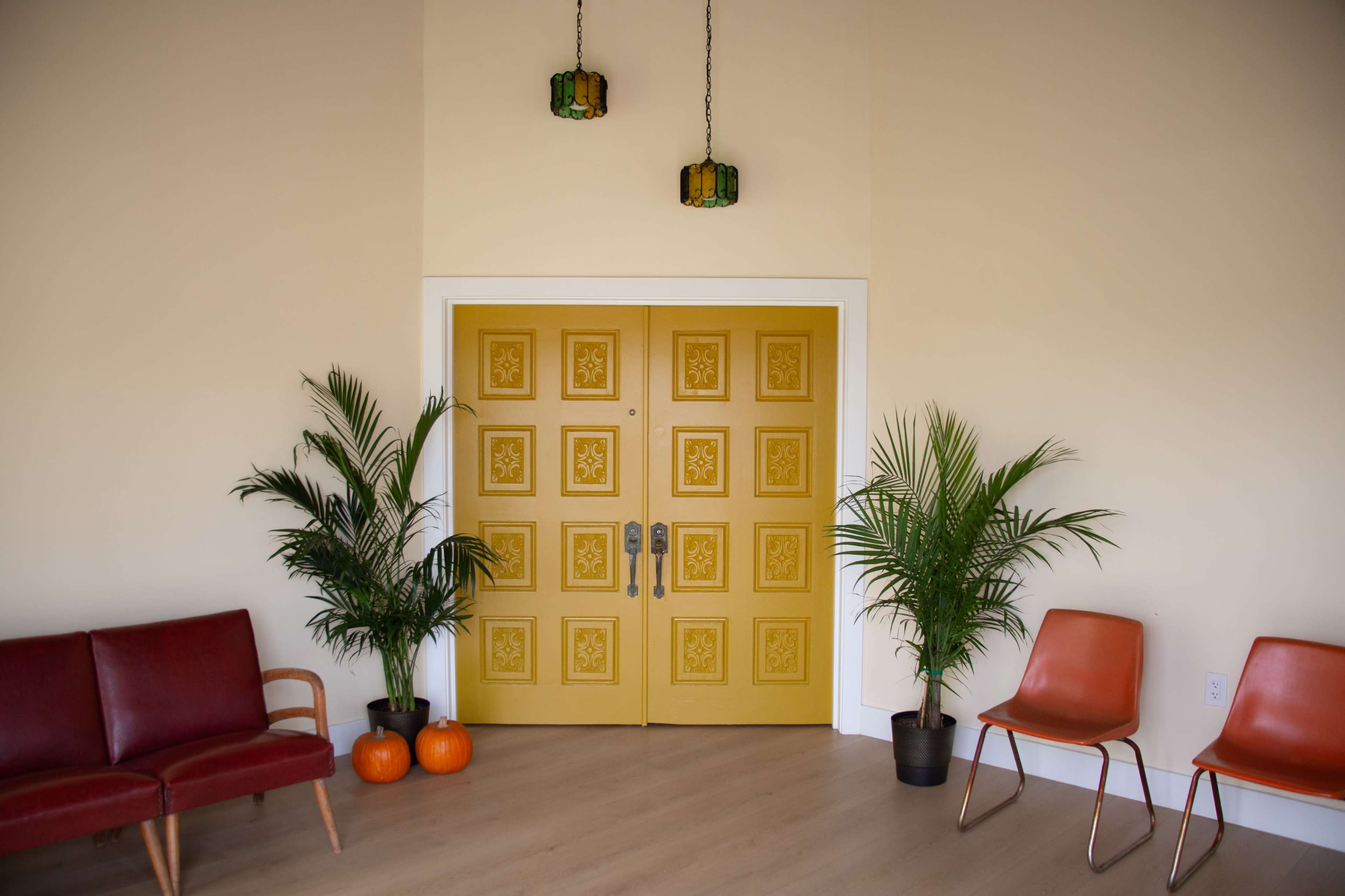 A bright yellow double door stands at the center of a room with minimalistic furniture, including a red sofa and orange chairs, flanked by potted plants and pumpkins.