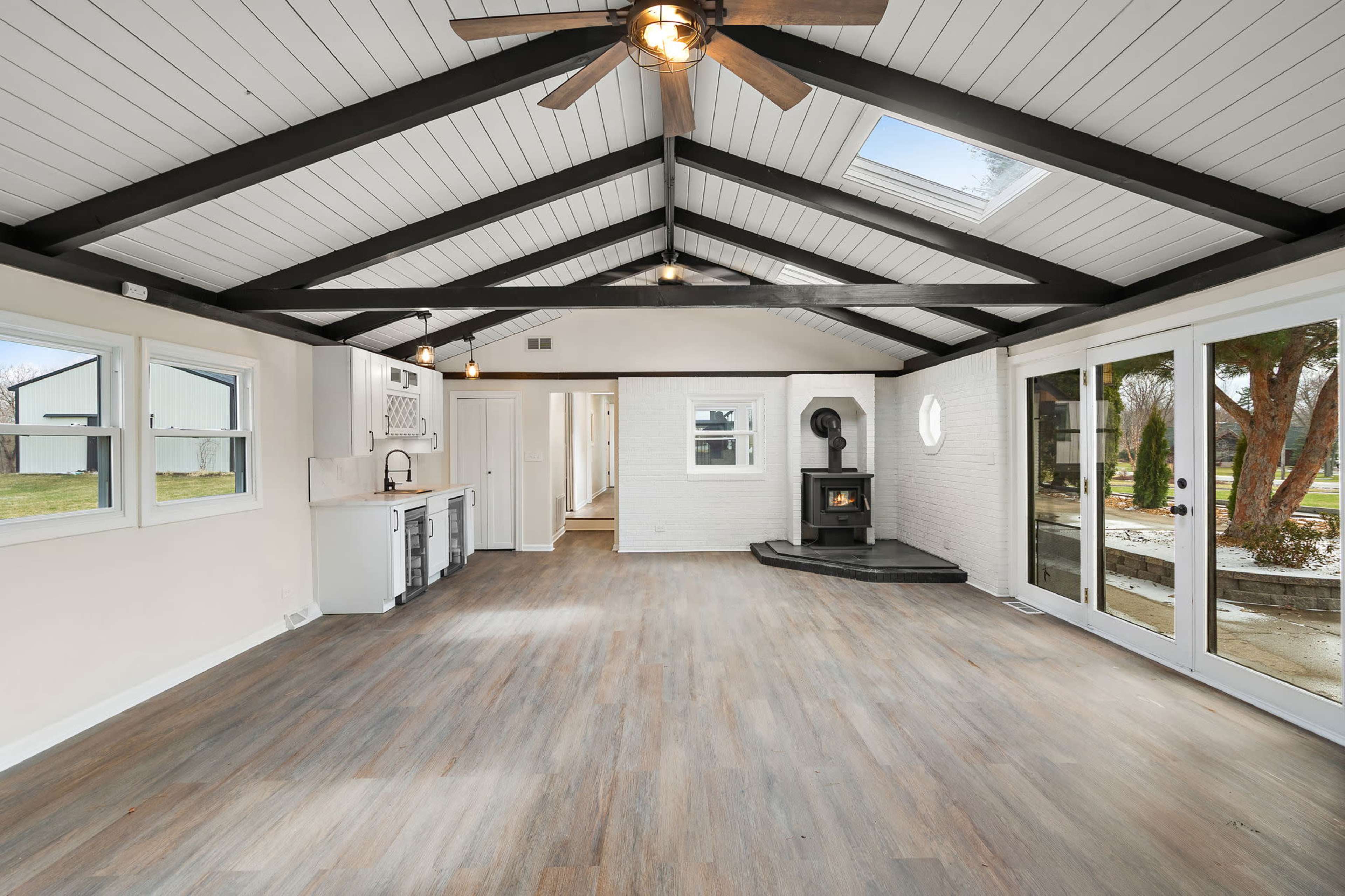 The image shows a spacious interior room with high vaulted ceilings, wooden beams, and large windows, featuring a corner stove and open access to an outdoor area.