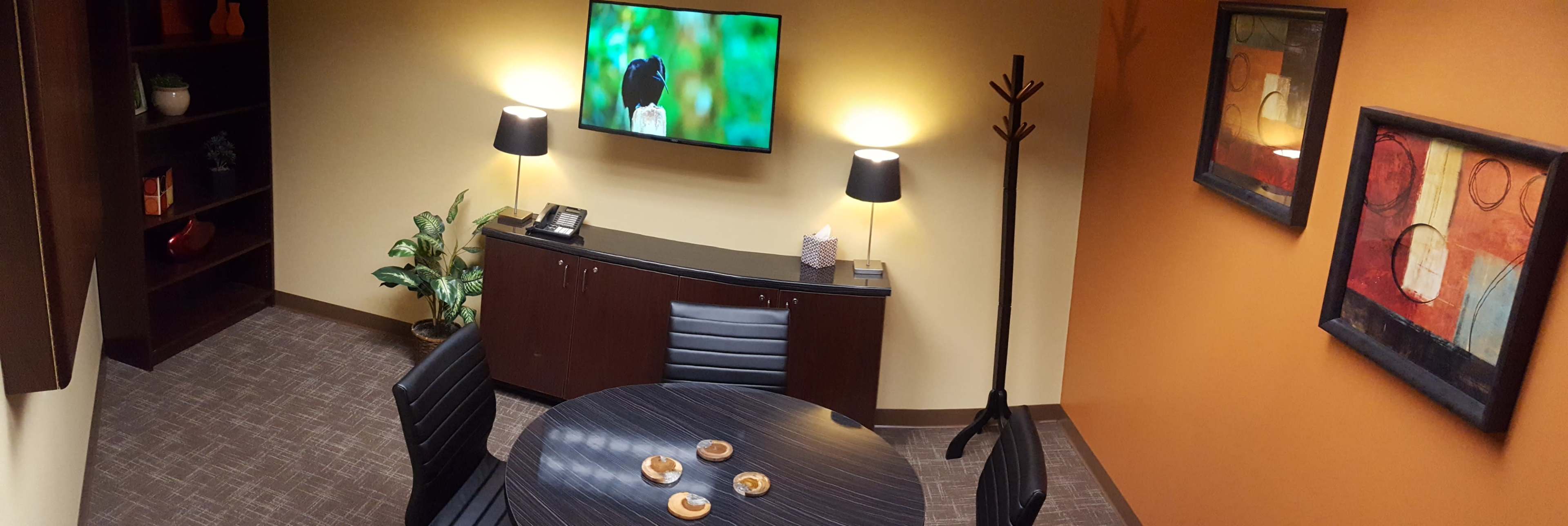 A small office meeting room features a round table with snacks, a television mounted on the wall, and decorations in warm colors.