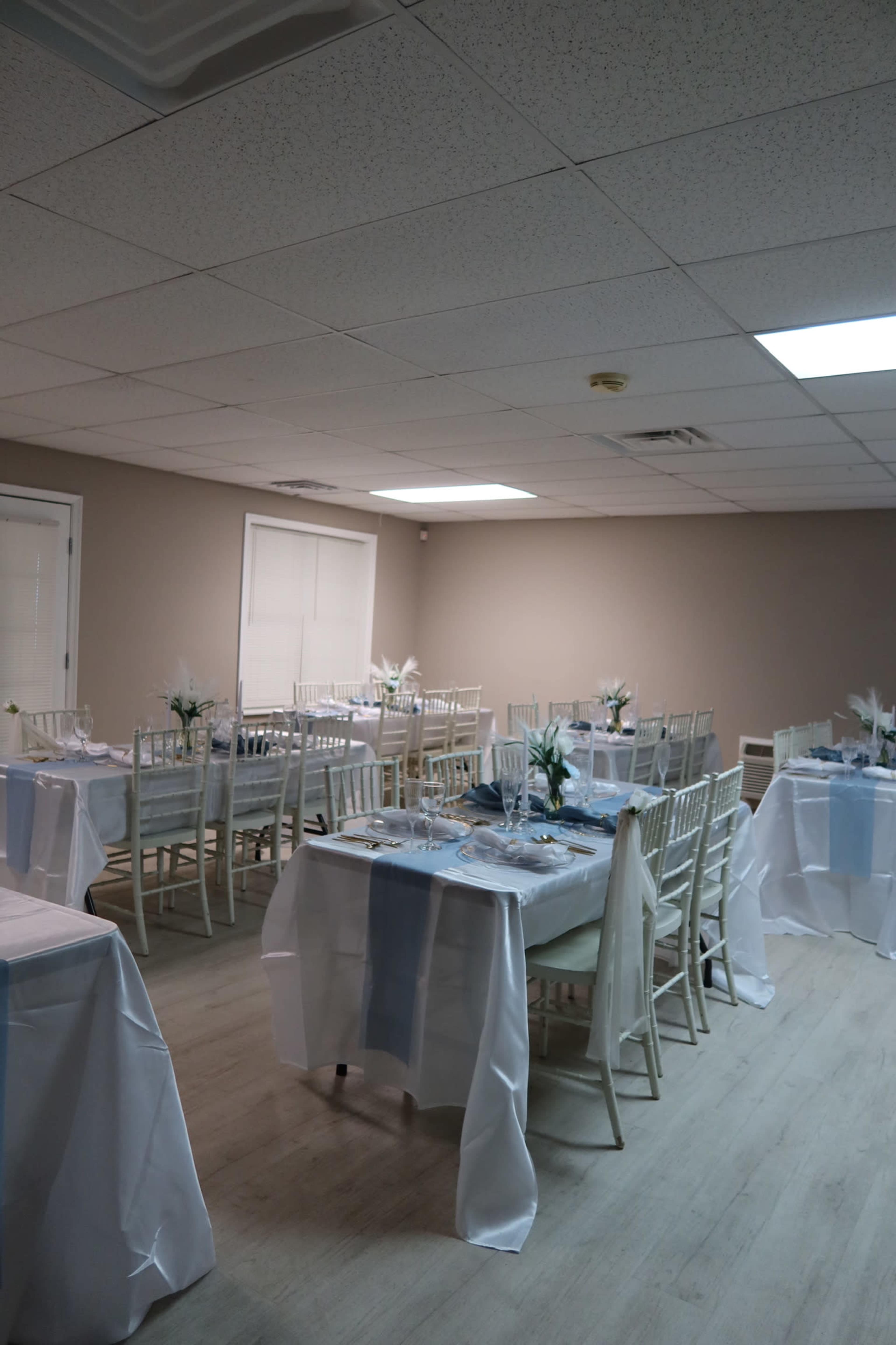 The image shows a banquet hall set with multiple tables covered in white tablecloths and arranged with chairs, each featuring decorative centerpieces.