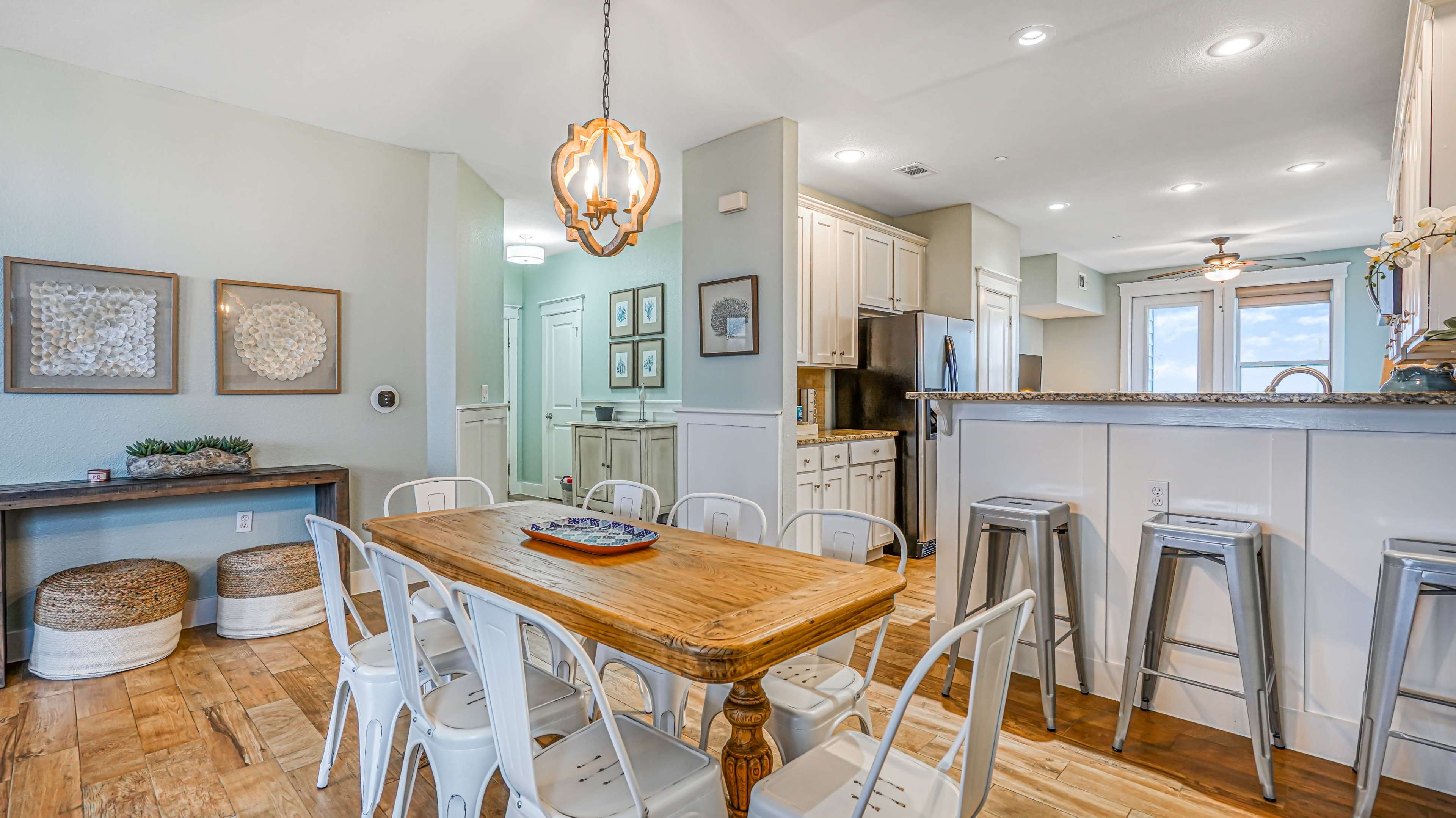 The image shows a dining area with a wooden table surrounded by white chairs, positioned near a kitchen with light-colored cabinets and a large window.