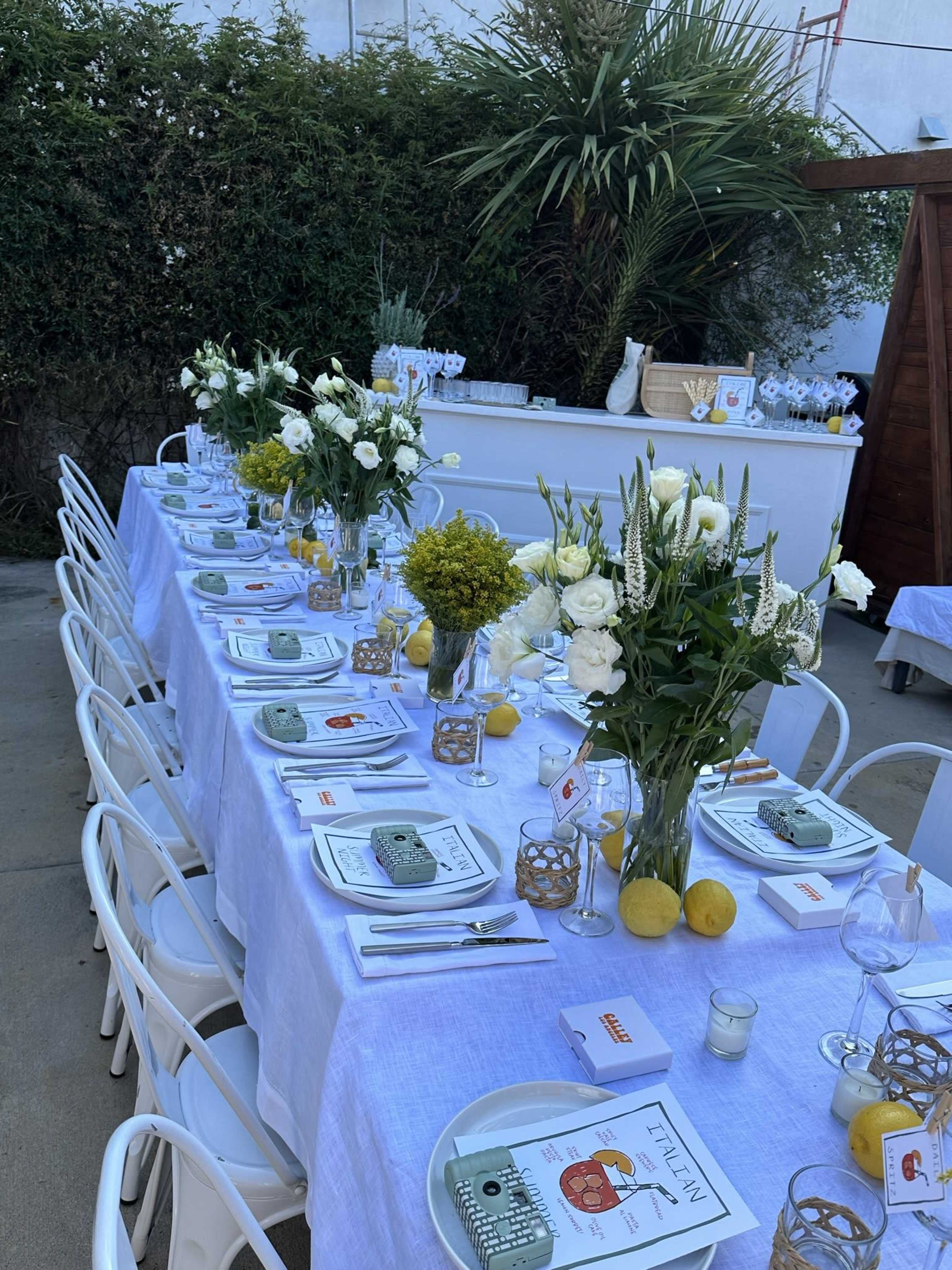 A long dining table is set outdoors with white tablecloths, floral centerpieces, and neatly arranged place settings, surrounded by lush greenery.