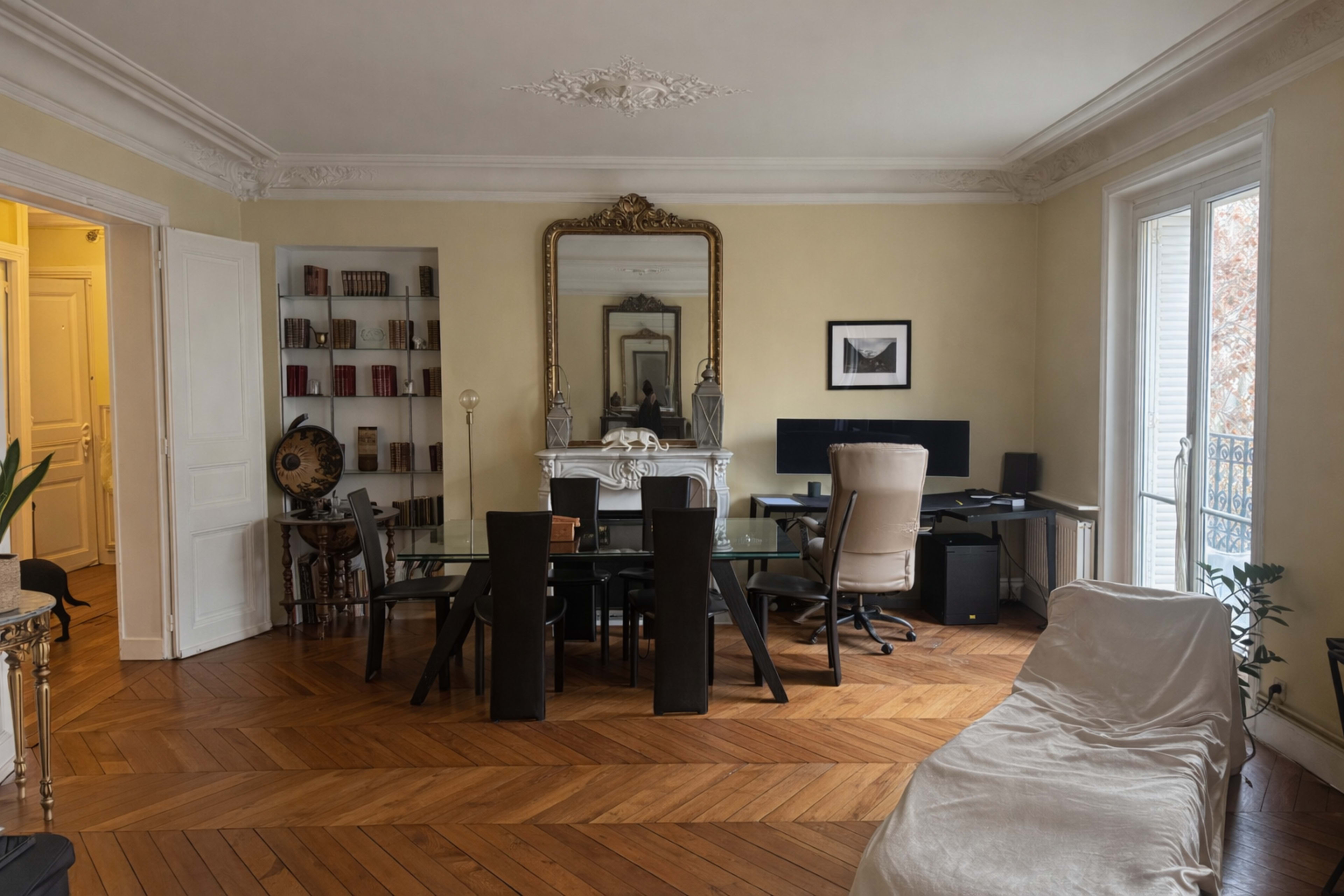 A spacious office features a large wooden table, black ergonomic chair, and a decorative mirror above a fireplace, with bookshelves and natural light from large windows.