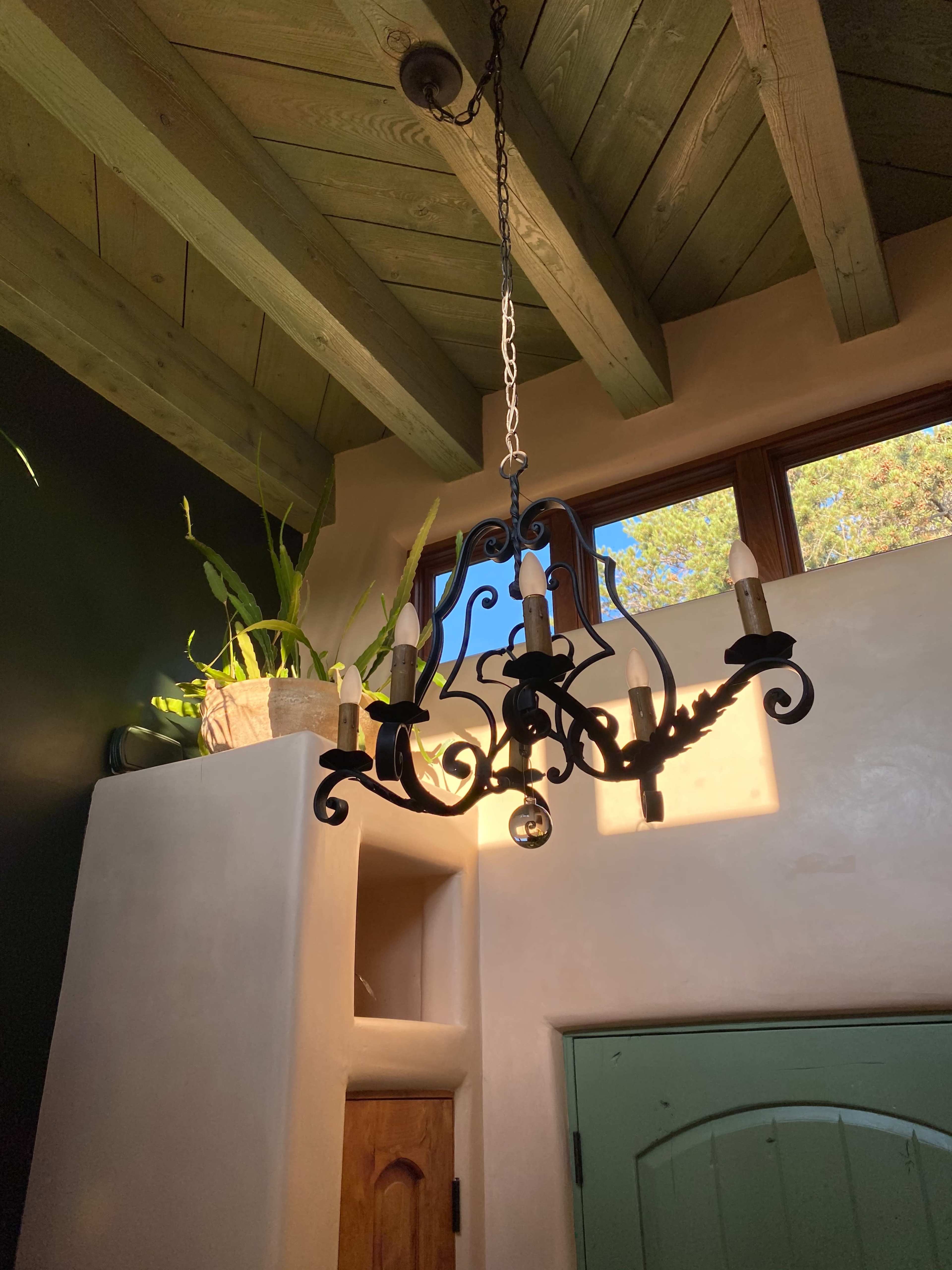 A wrought iron chandelier hangs from a wooden beam ceiling, illuminated by sunlight coming through a window.
