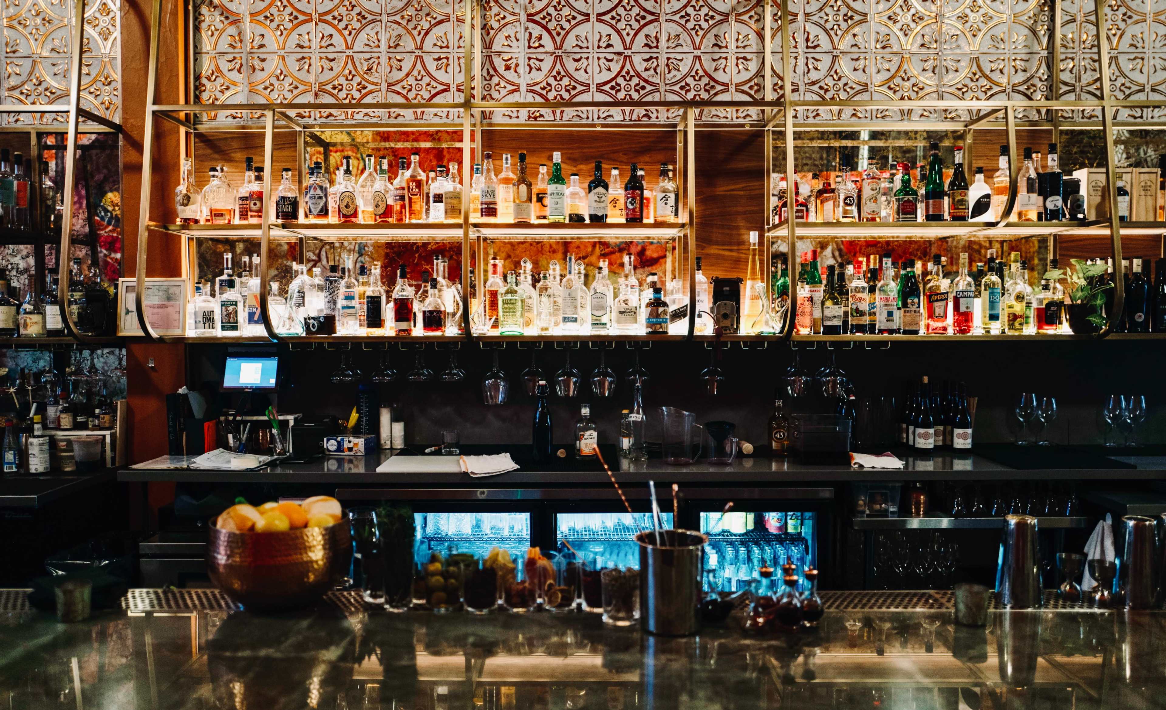 A well-organized bar features illuminated shelves filled with various bottles of alcohol and a polished countertop with glassware and mixers.