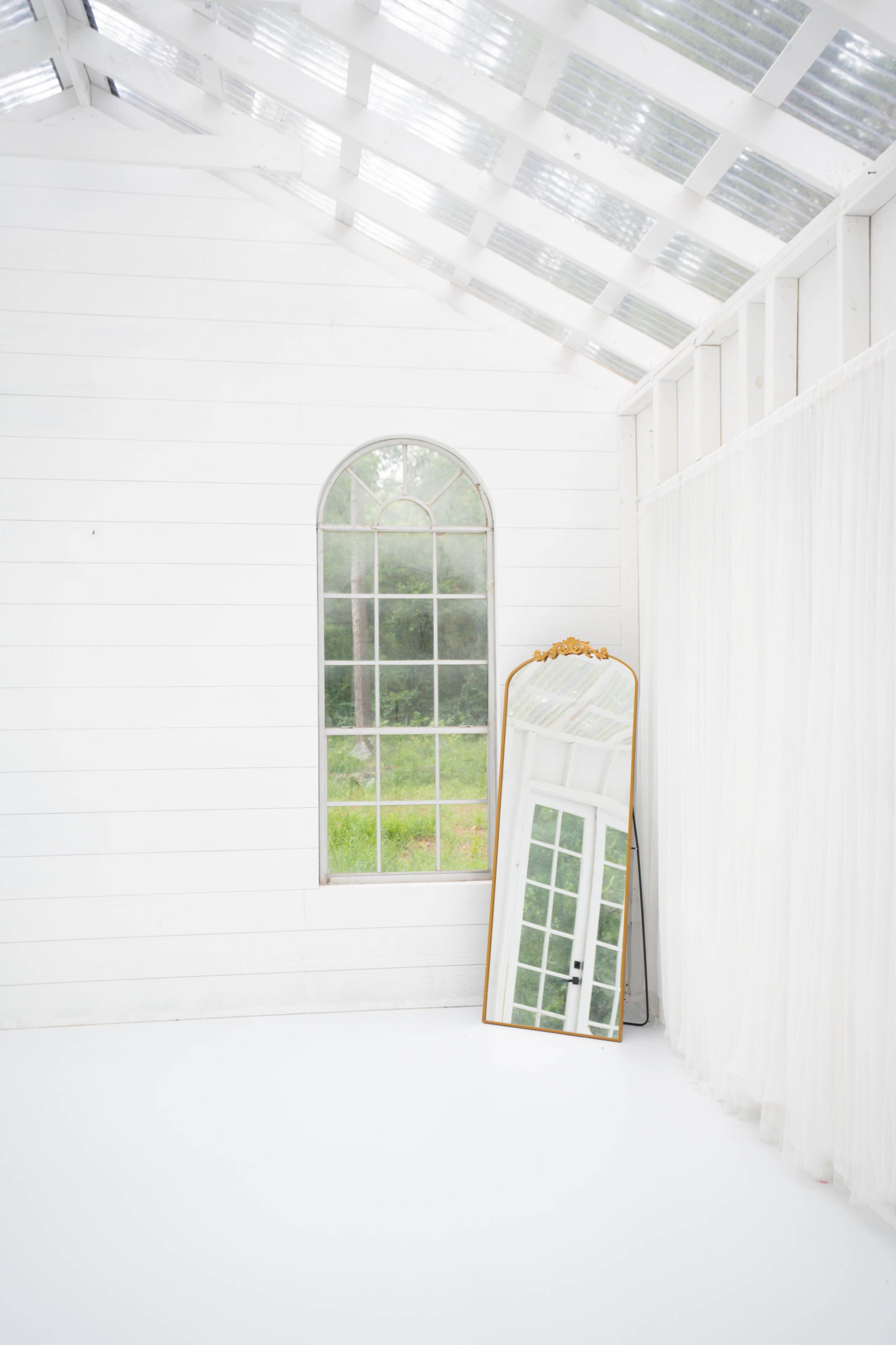 The image shows a bright, minimalist room with white wooden walls, a large arched window, and a tall mirror reflecting the window.