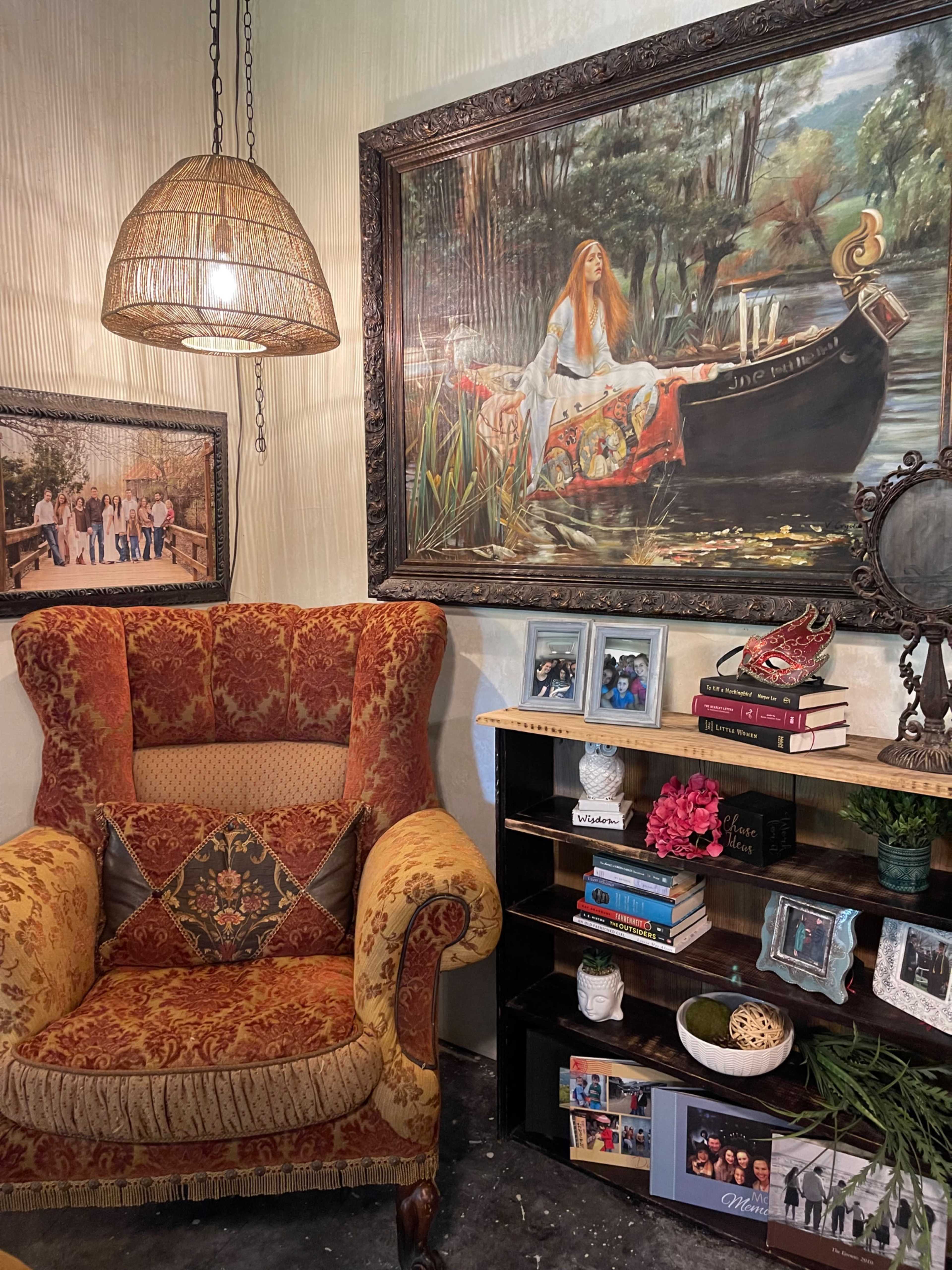 The image features a cozy corner with an ornate armchair beside a wooden shelf displaying books and decorative items, while a large painting of a woman on a boat hangs on the wall.