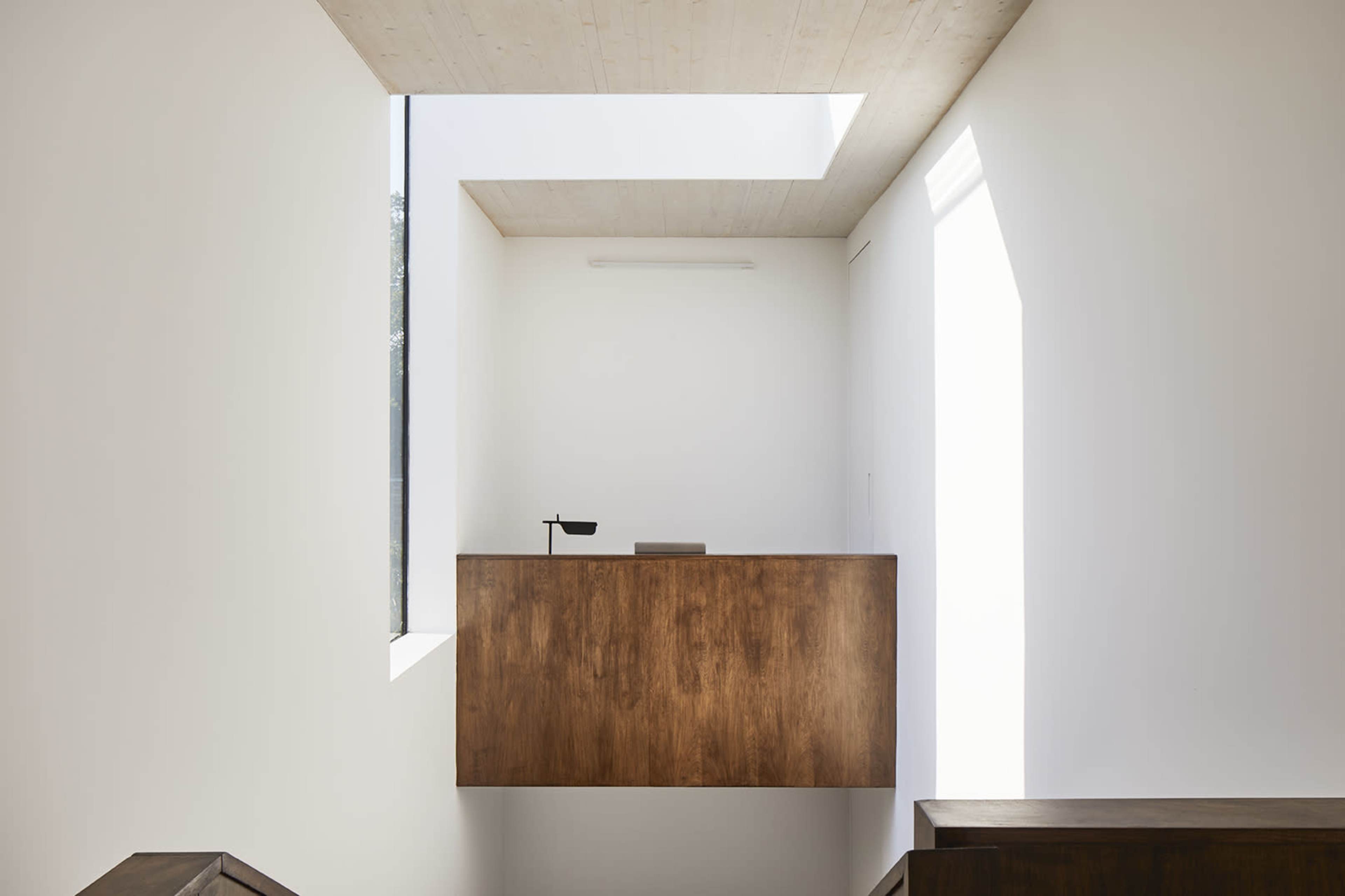 A wooden reception desk is positioned against a white wall in a well-lit, modern interior space featuring a skylight.