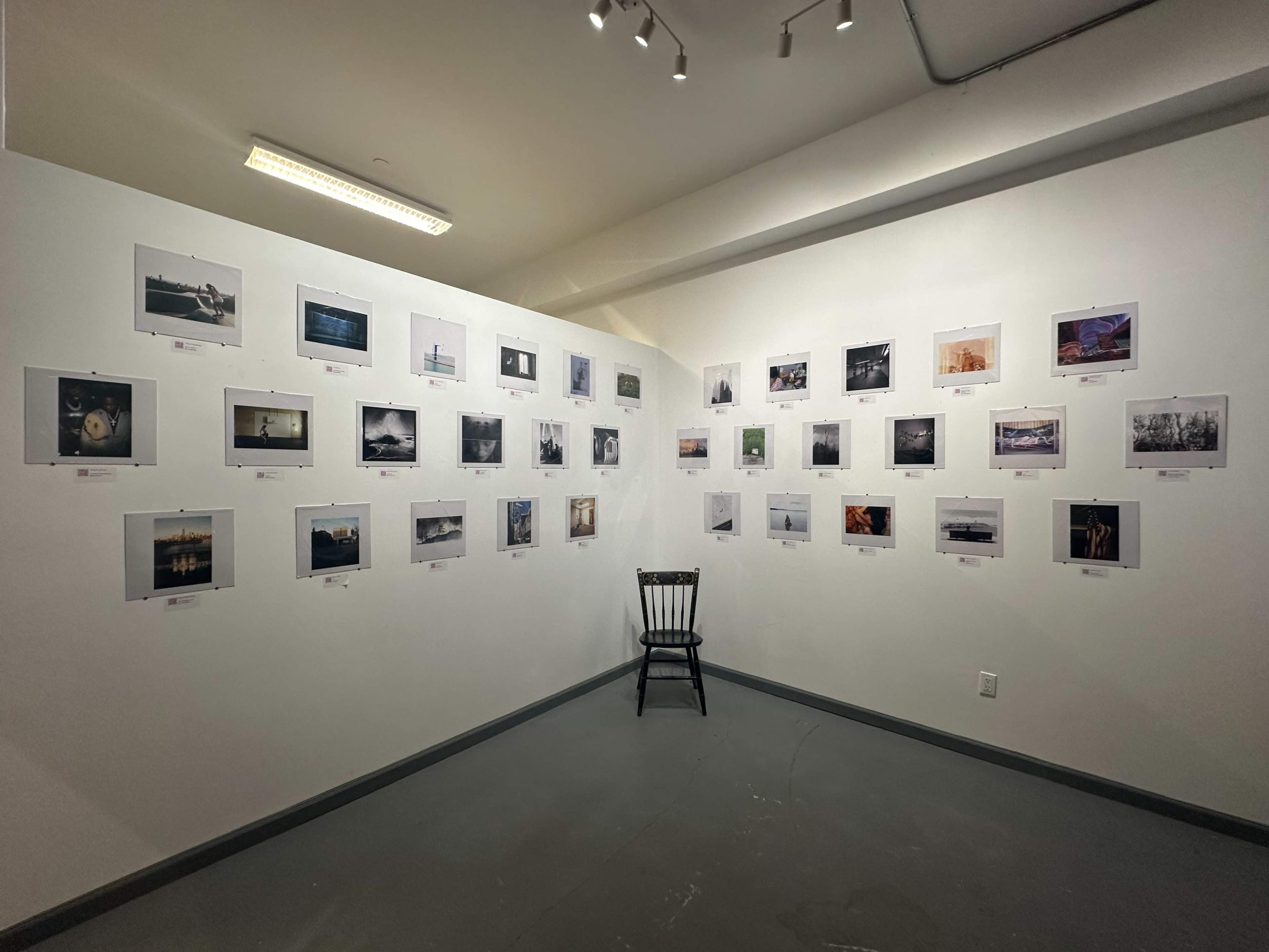 The image shows a gallery exhibition featuring a grid of framed photographs displayed on two adjacent walls, with a single black chair positioned in the center of the room.
