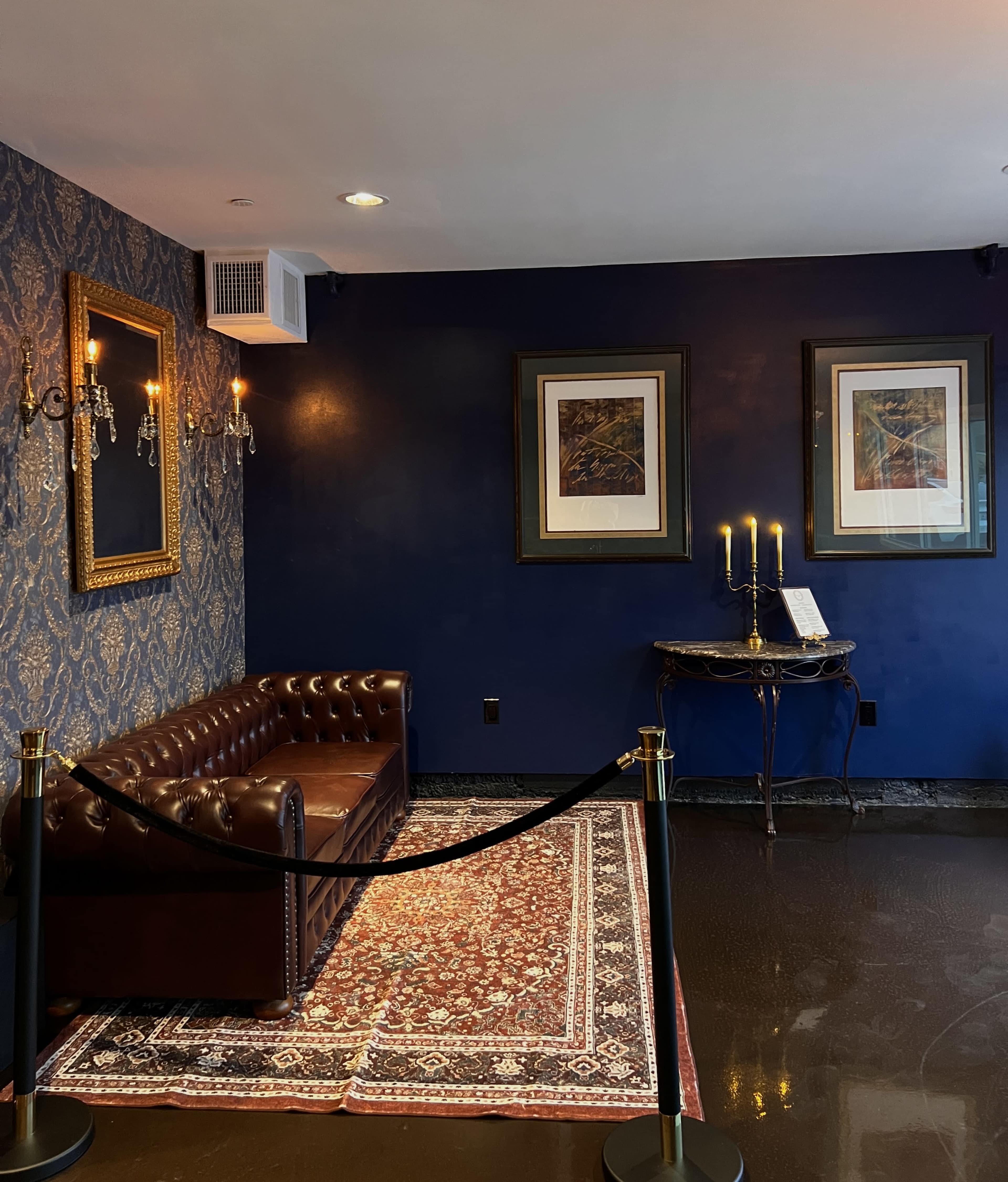 The image shows a cozy corner of a room featuring a brown leather couch, a patterned area rug, framed artwork on the wall, and a decorative table with candles.