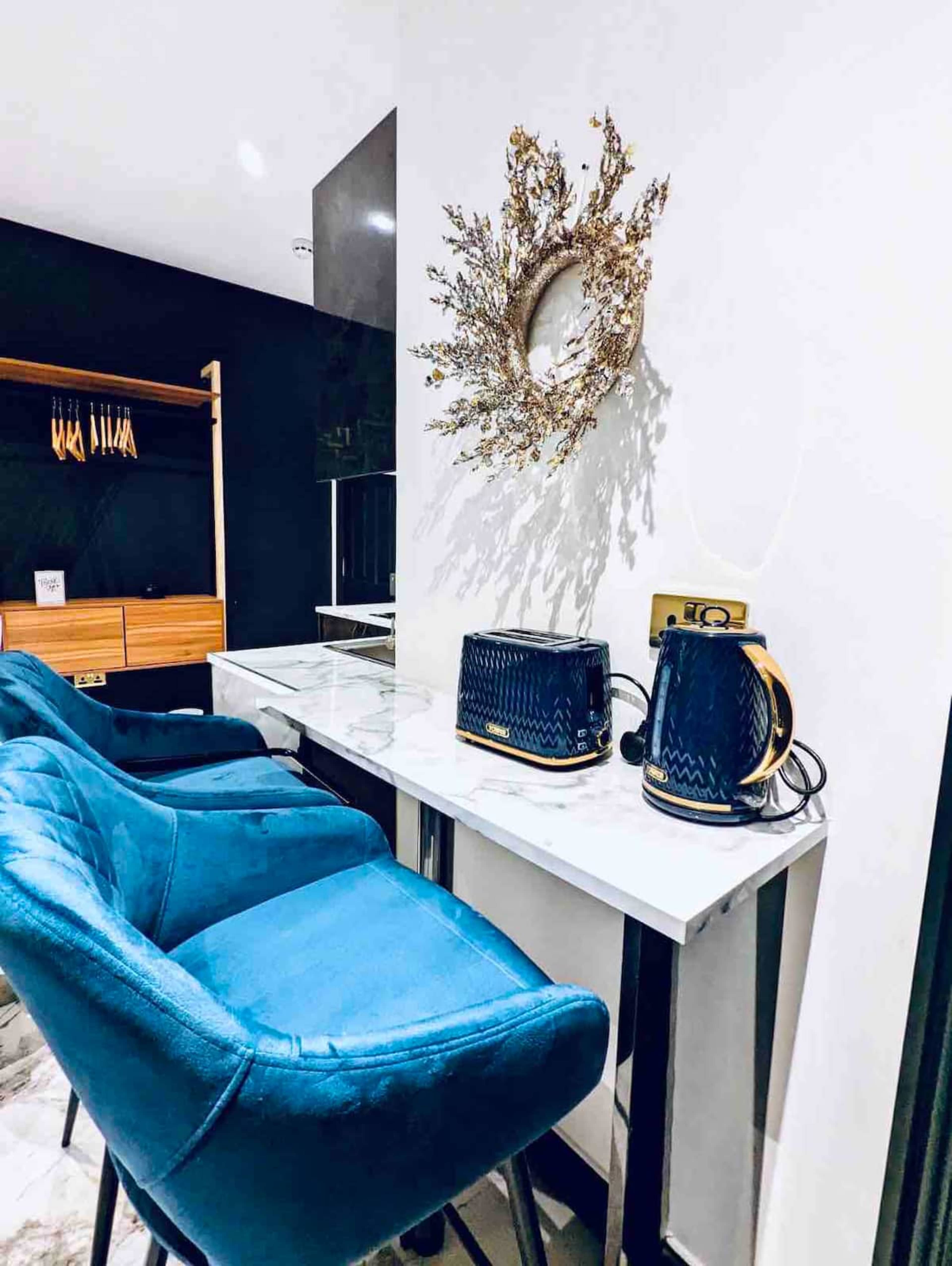 The image shows a modern kitchen space with two blue velvet chairs, a glass table, and a decorative wall piece above a countertop featuring a kettle and toaster.