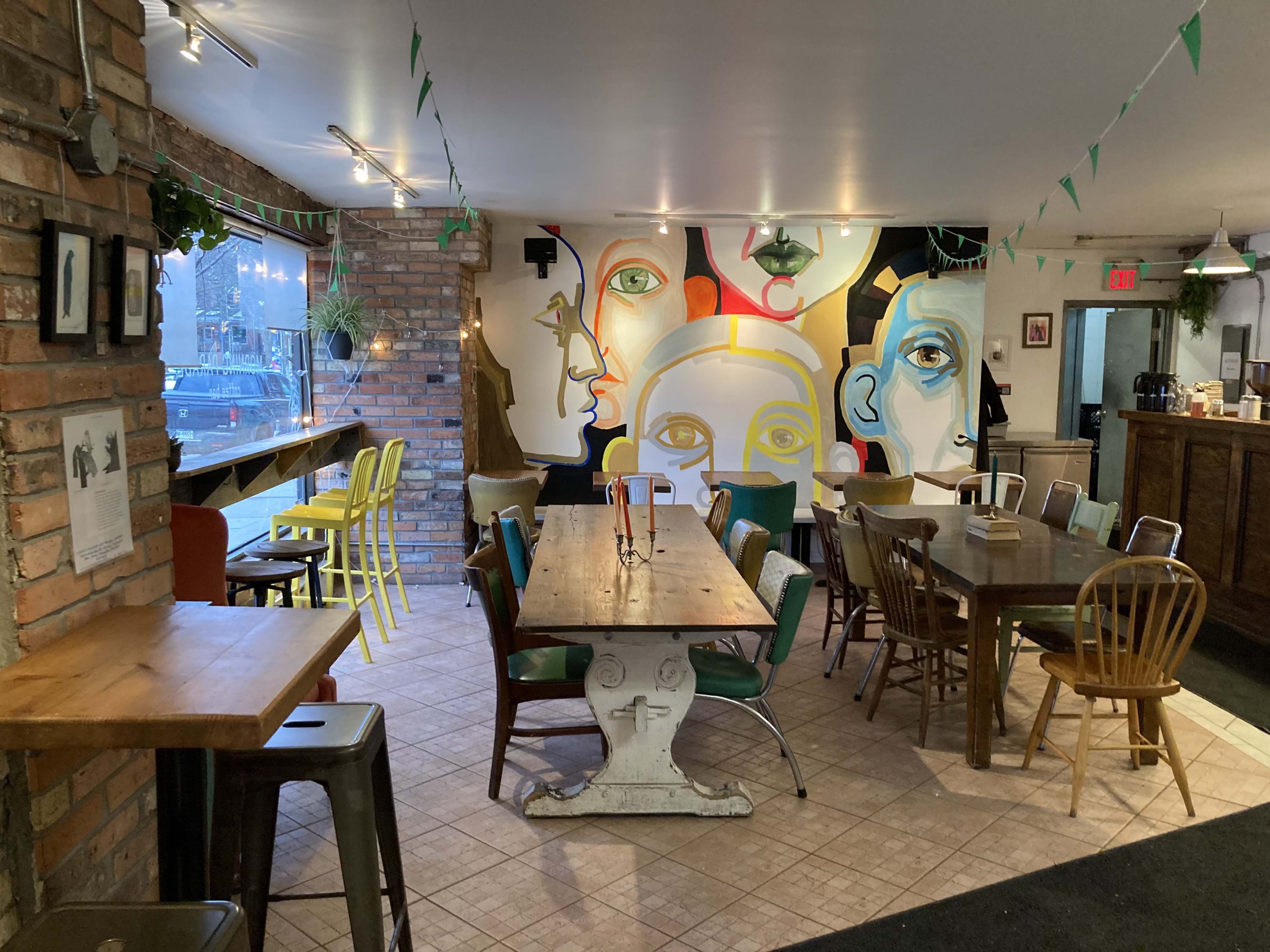The image shows a café interior featuring a mix of tables and chairs along with a vibrant mural of faces on one wall.