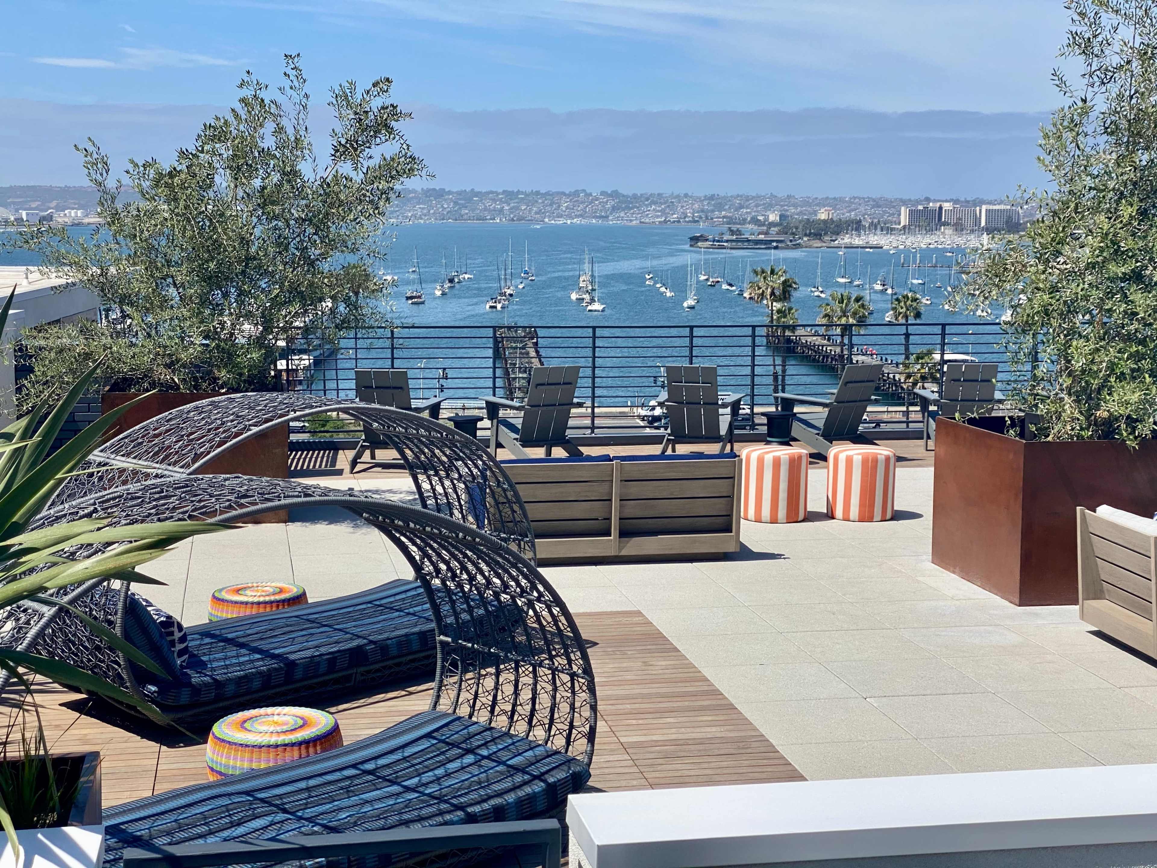 The image shows a rooftop terrace with lounge chairs and decorative seating overlooking a marina filled with boats and a coastal landscape.