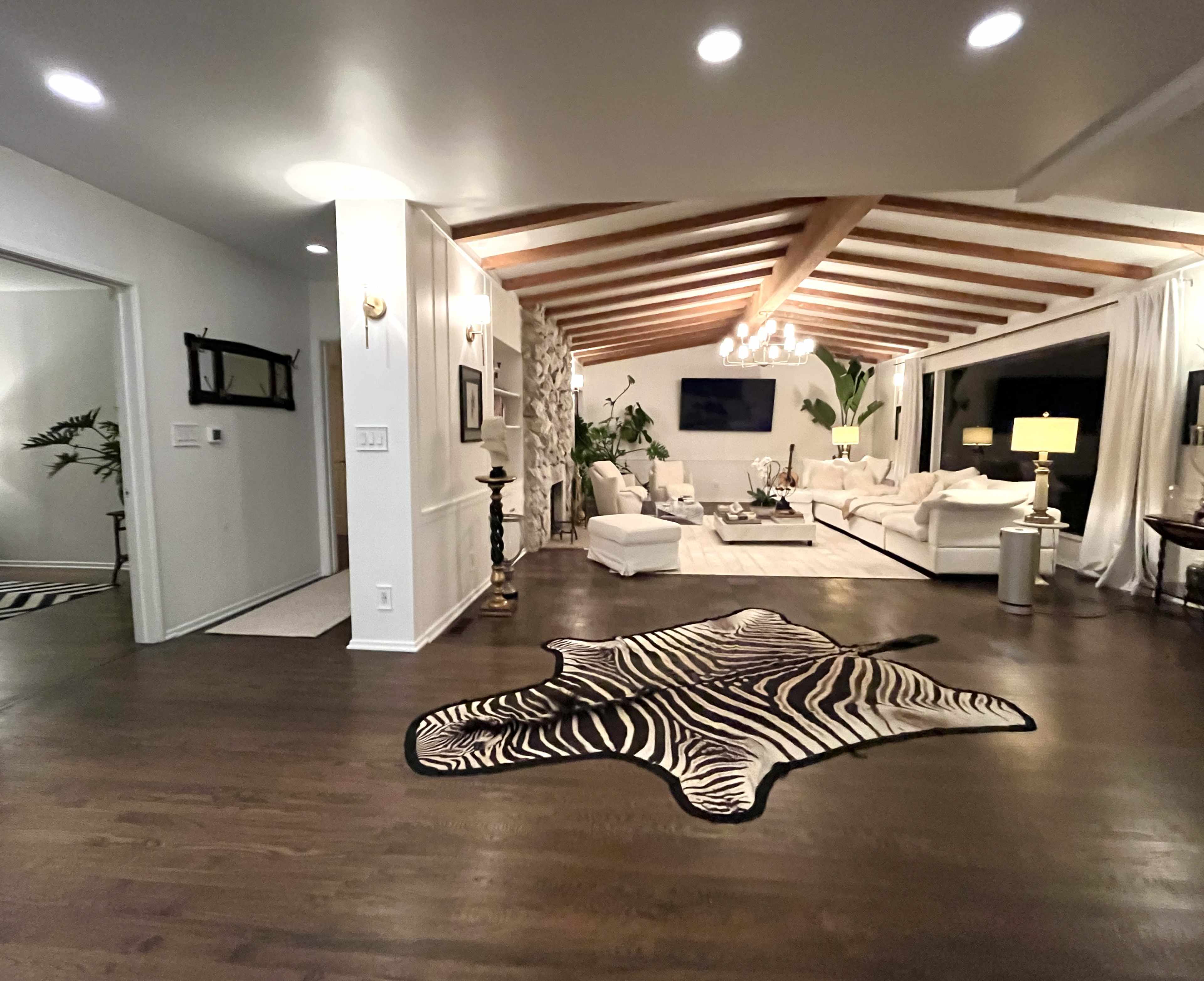 The image shows a modern living room featuring a white sectional sofa, decorative plants, a zebra-print rug, and wooden beams on the ceiling.