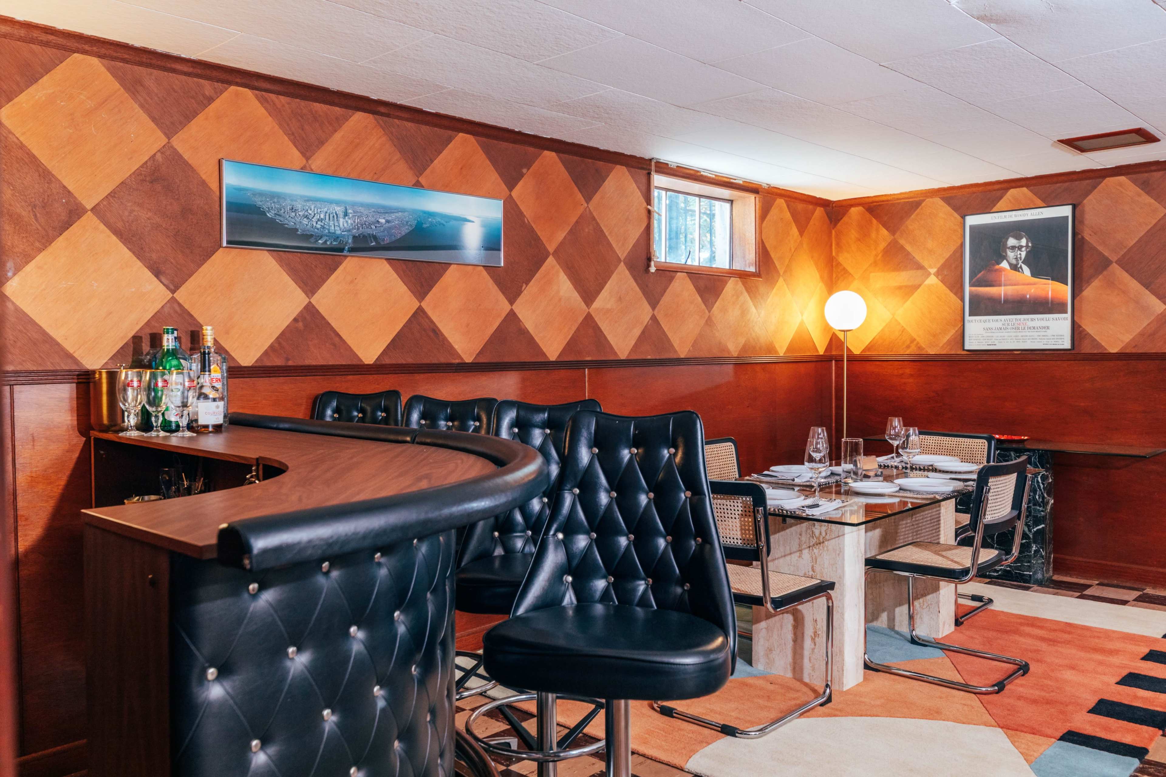 The image shows a bar area with black upholstered stools and a dining table set in a room featuring a diamond-patterned wall design and various decorative elements.