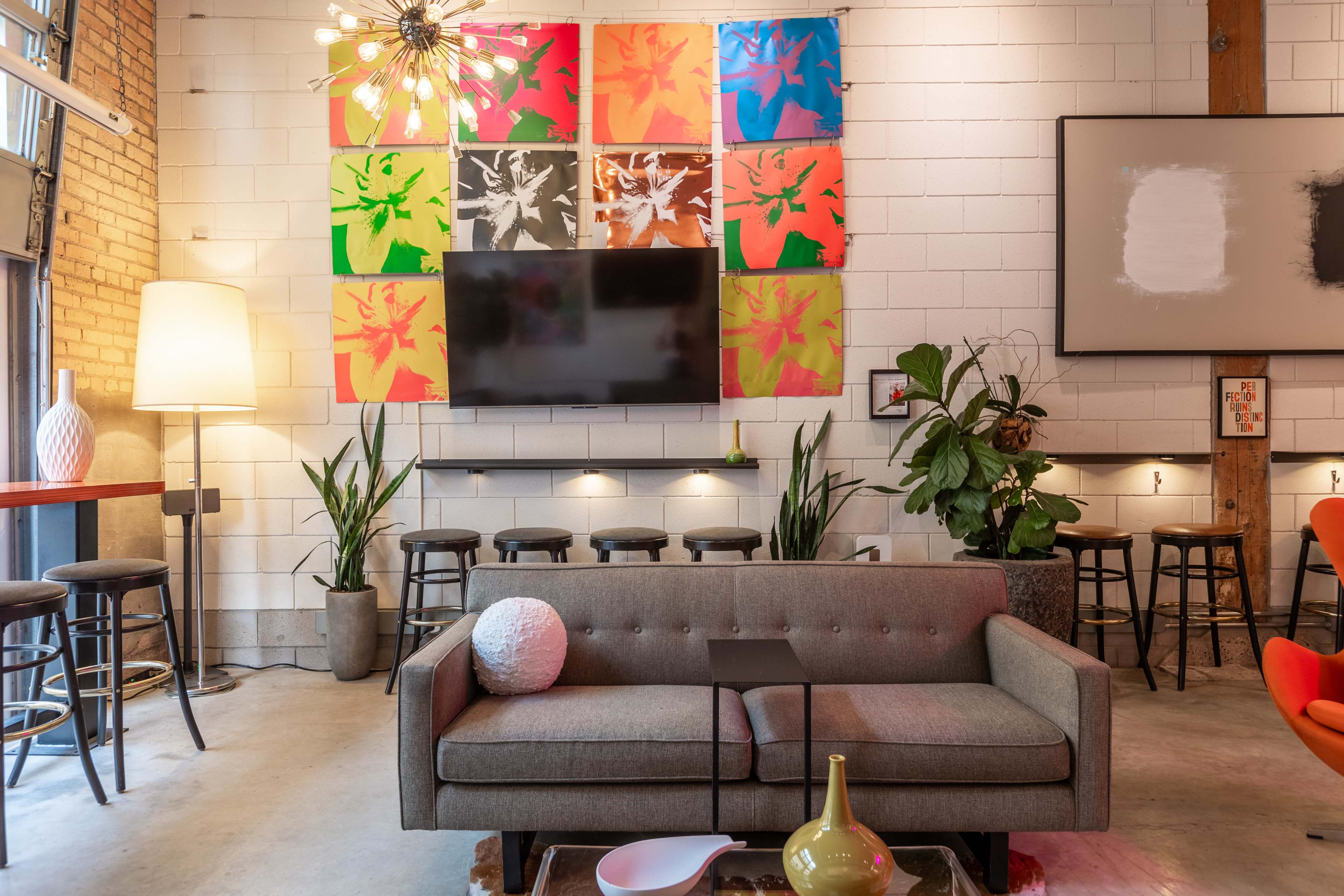 The image shows a modern lounge area featuring a gray sofa, a large wall-mounted television, colorful artwork, bar stools, and potted plants against a backdrop of exposed brick.