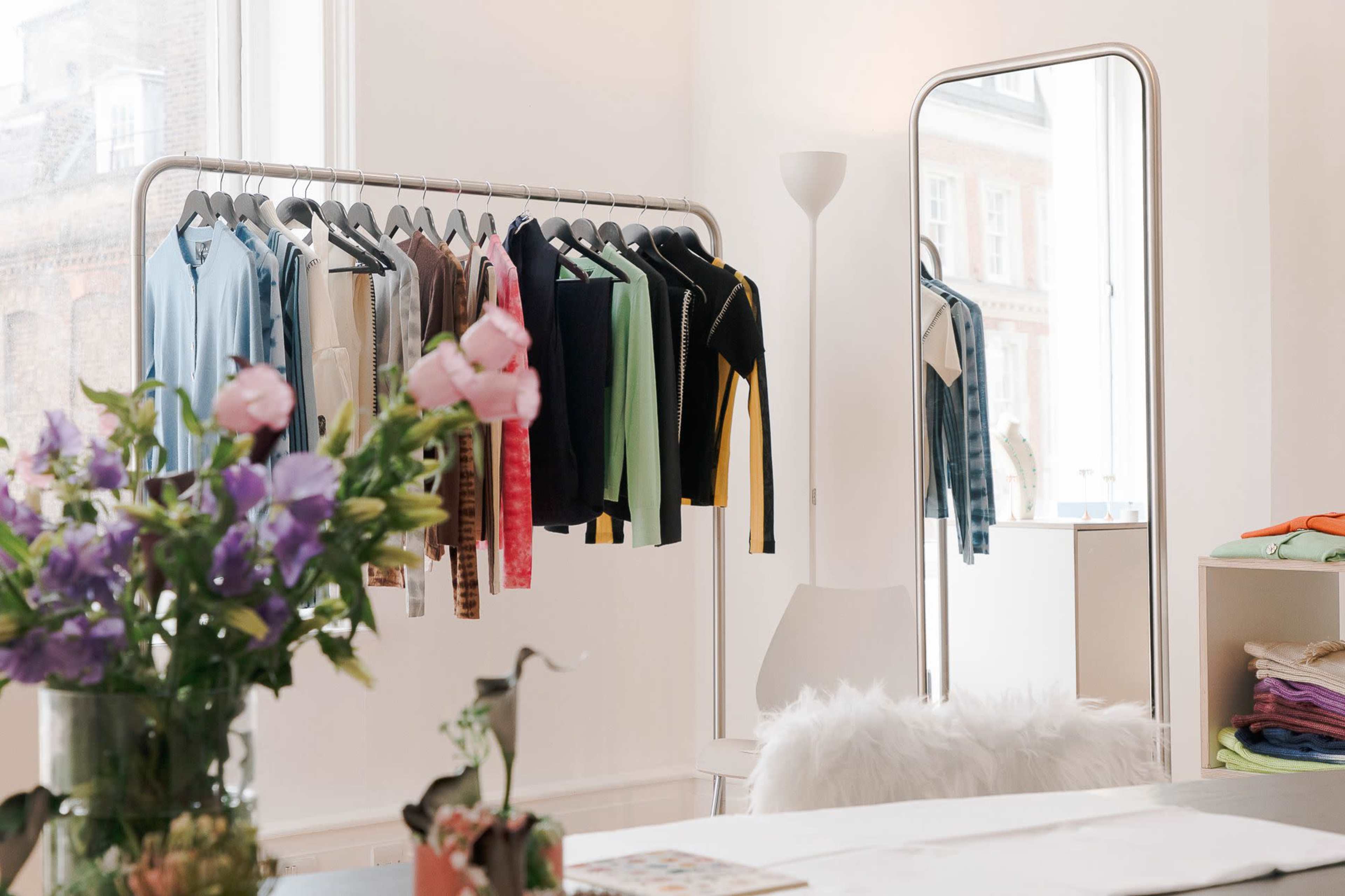 A clothing rack holds a variety of shirts in a well-lit room with a mirror and a table adorned with flowers.