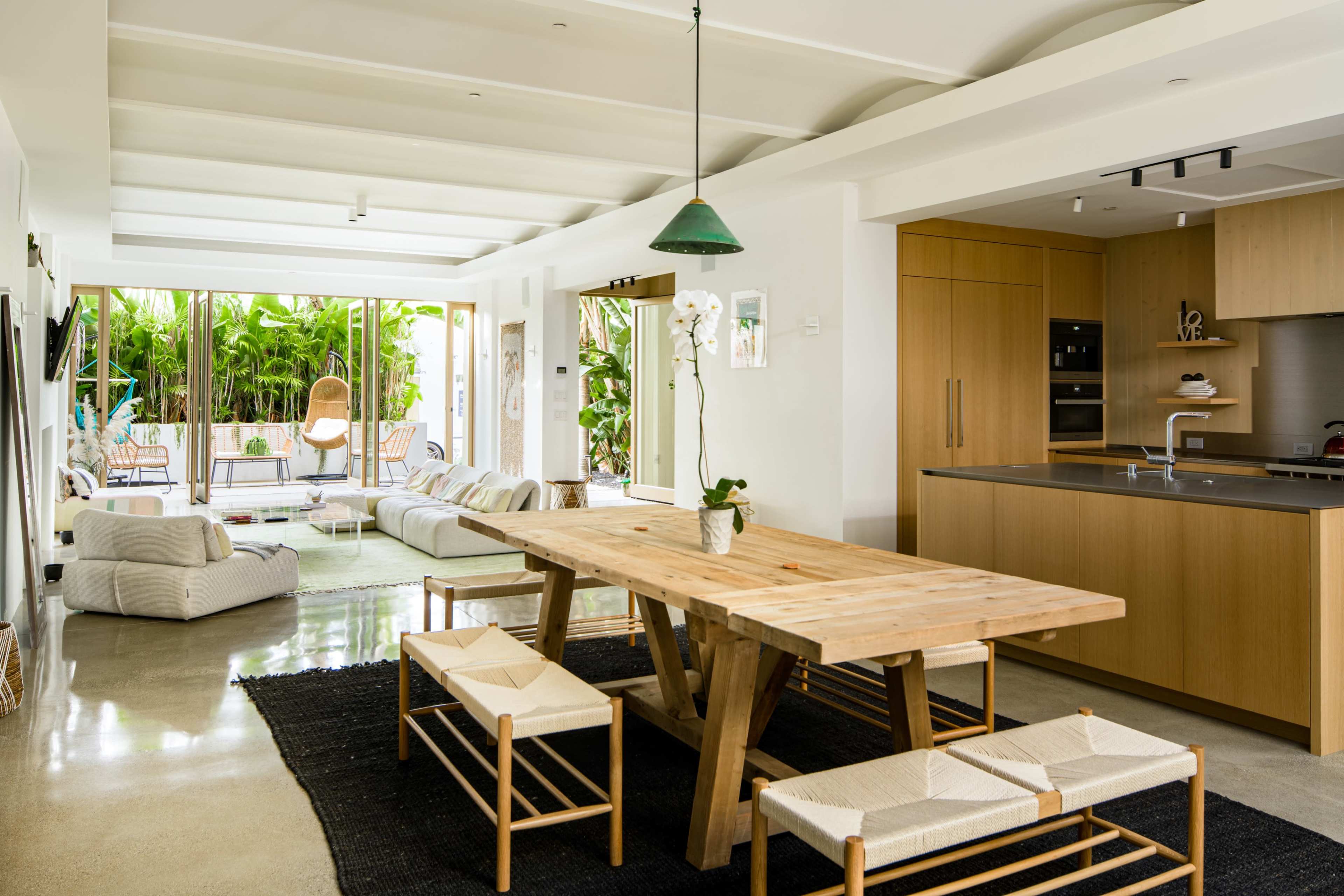 A spacious living area features a wooden dining table and light-colored sofas, with large glass doors opening to a patio surrounded by greenery.