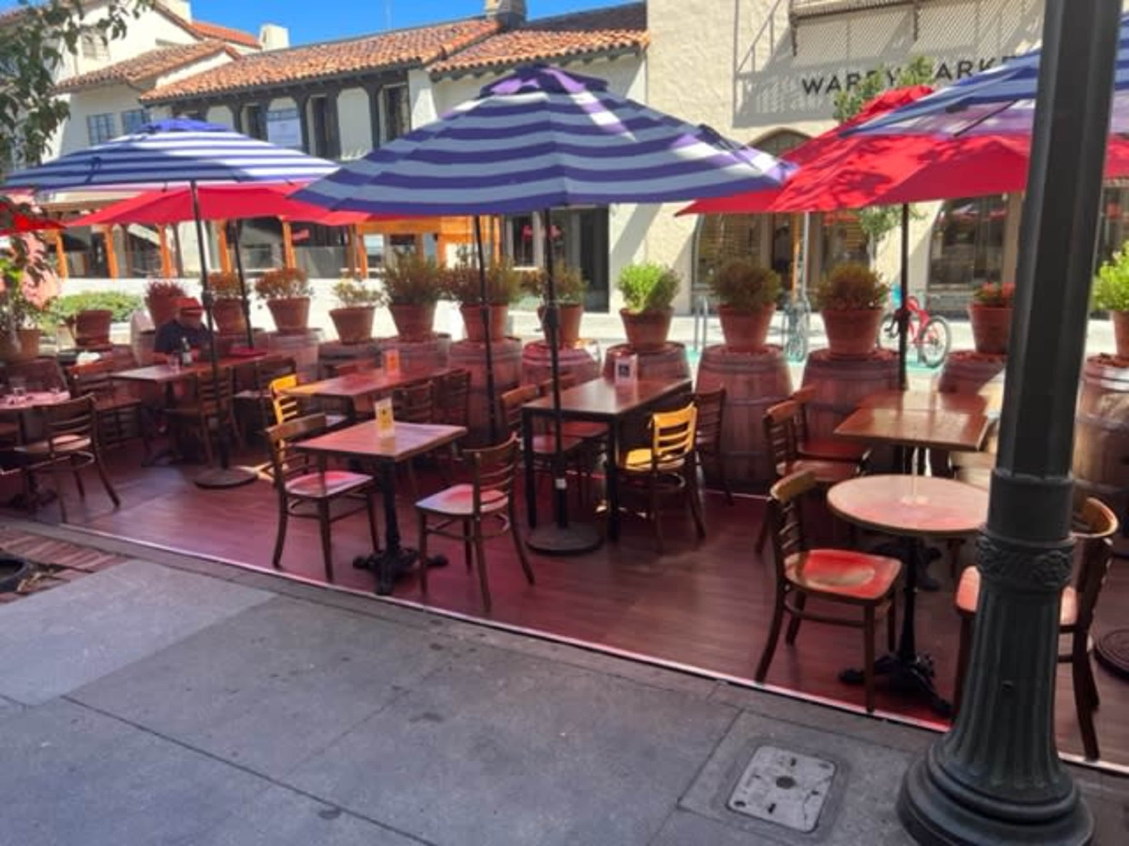 A row of outdoor tables and chairs, shaded by striped umbrellas, is set up outside a restaurant with potted plants along the perimeter.