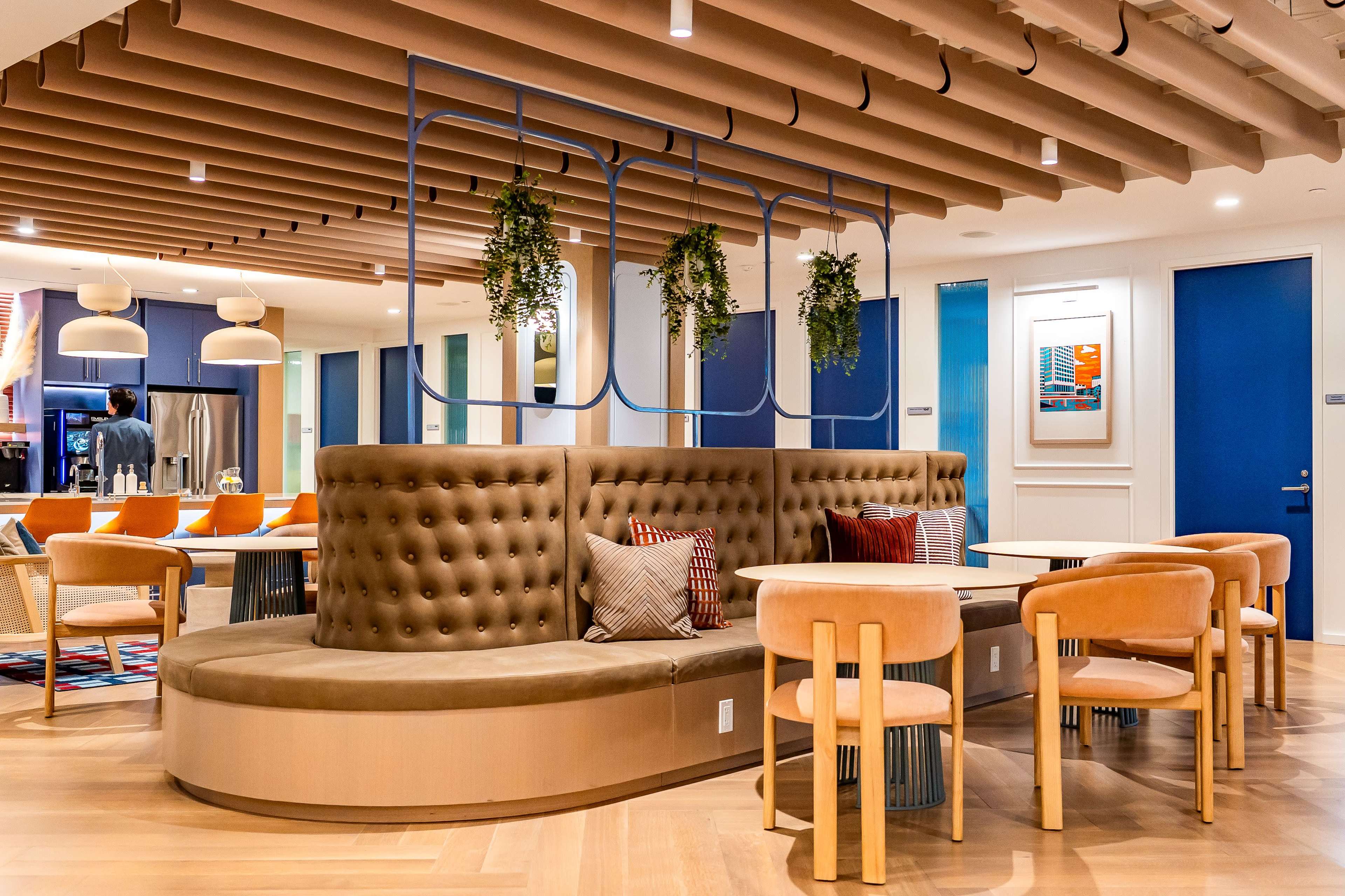 The image shows a modern lounge area featuring a circular upholstered seating arrangement with decorative pillows, surrounded by wooden tables and chairs, and accented by hanging plants and contemporary lighting fixtures.
