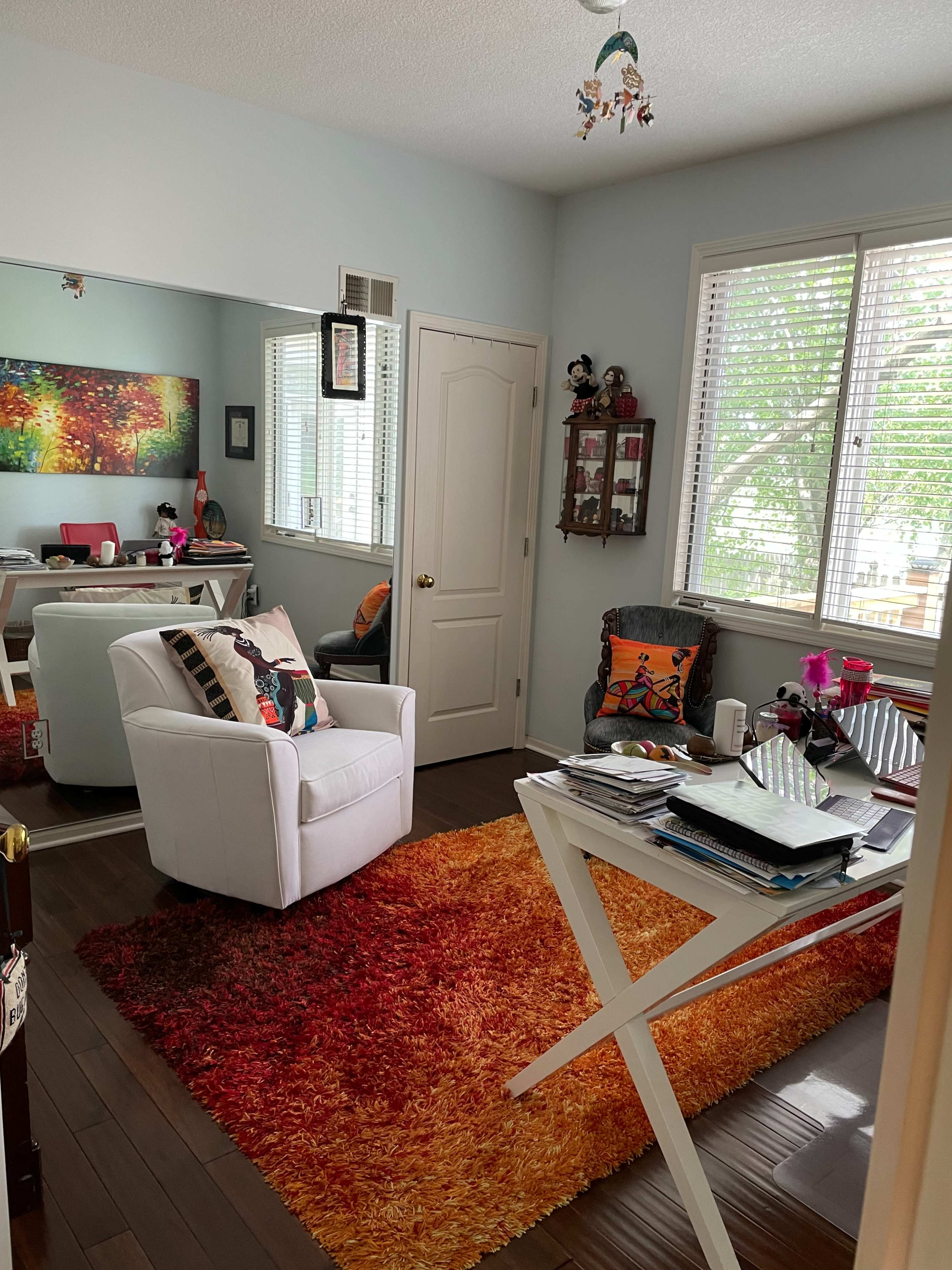 The image shows a brightly lit room with modern furnishings, including a white chair, a colorful area rug, a desk covered with papers, and windows letting in natural light.