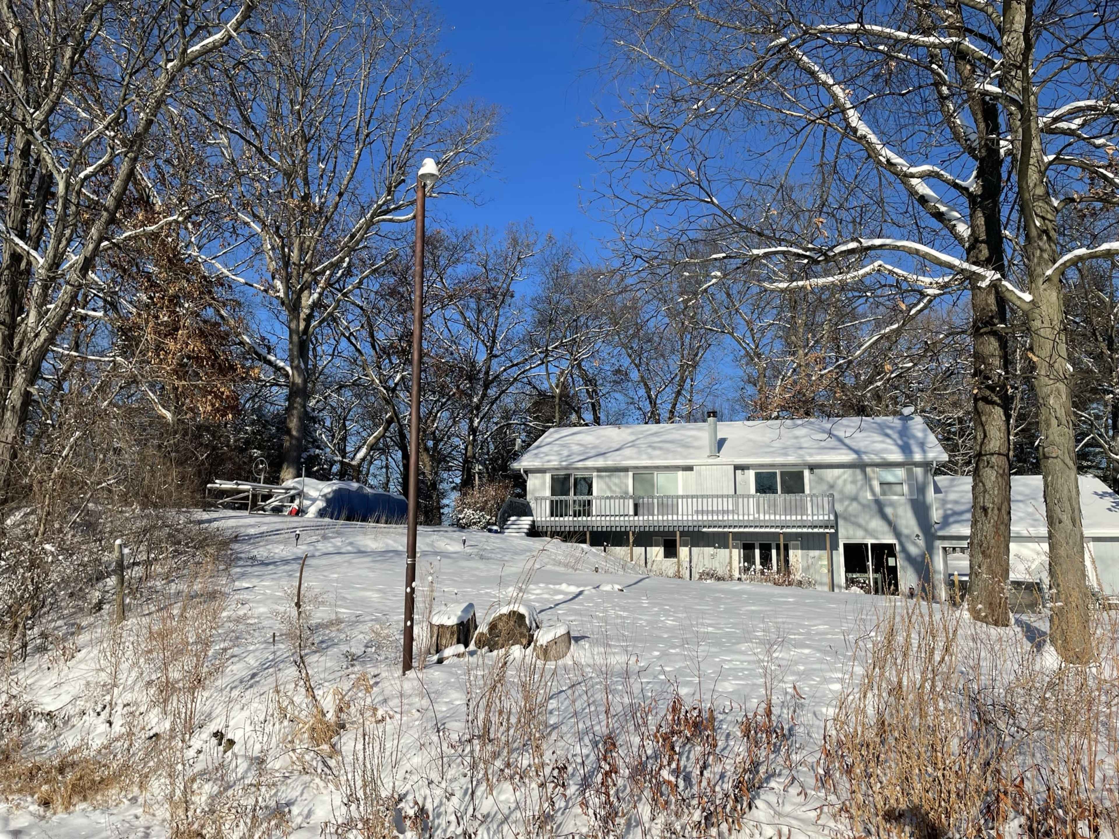 A two-story house stands on a snowy hillside surrounded by leafless trees under a clear blue sky.