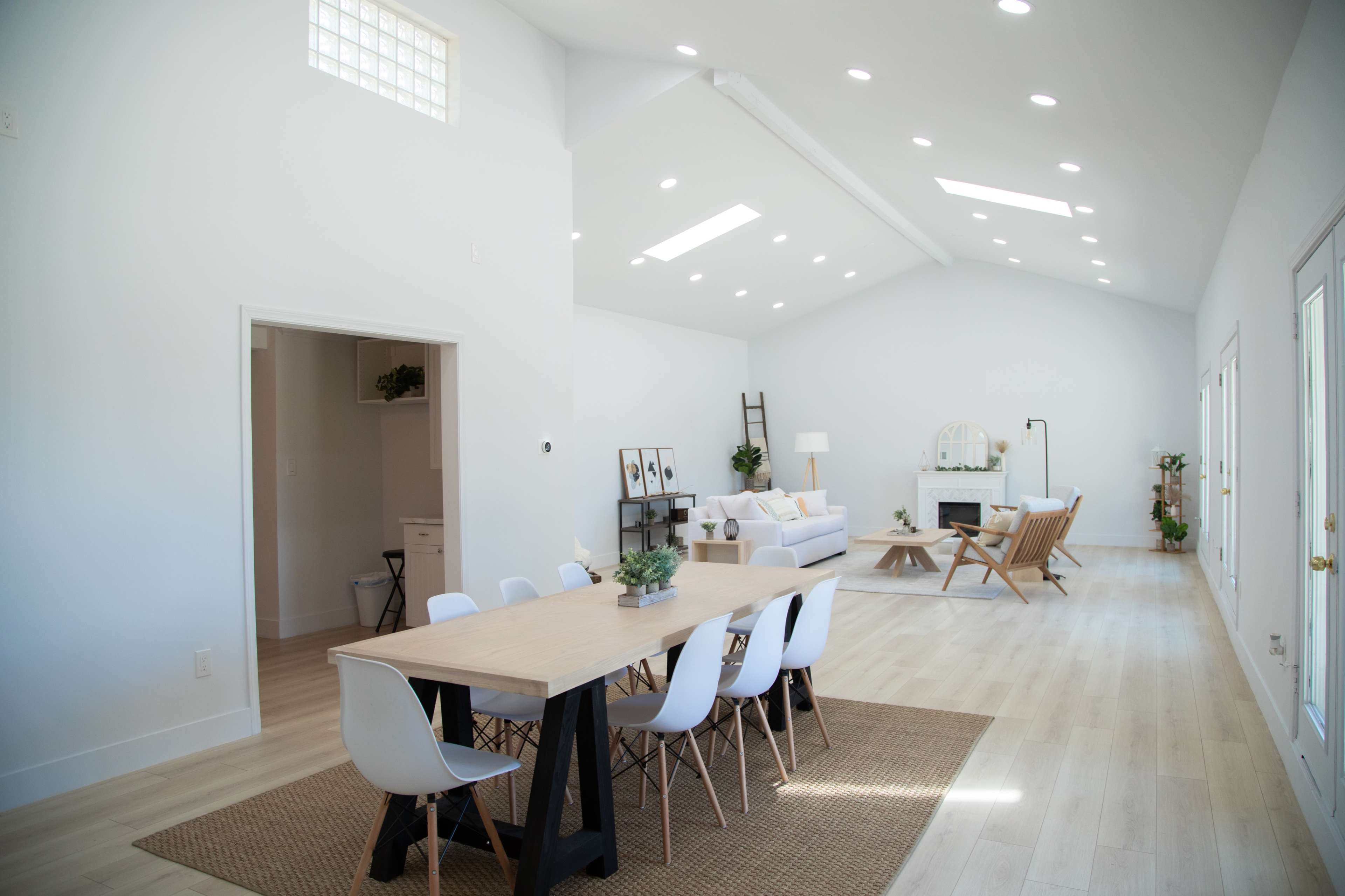 The image shows a spacious, brightly lit interior of a modern living area and dining space with a wooden table surrounded by white chairs, a seating area with a sofa, and large windows.