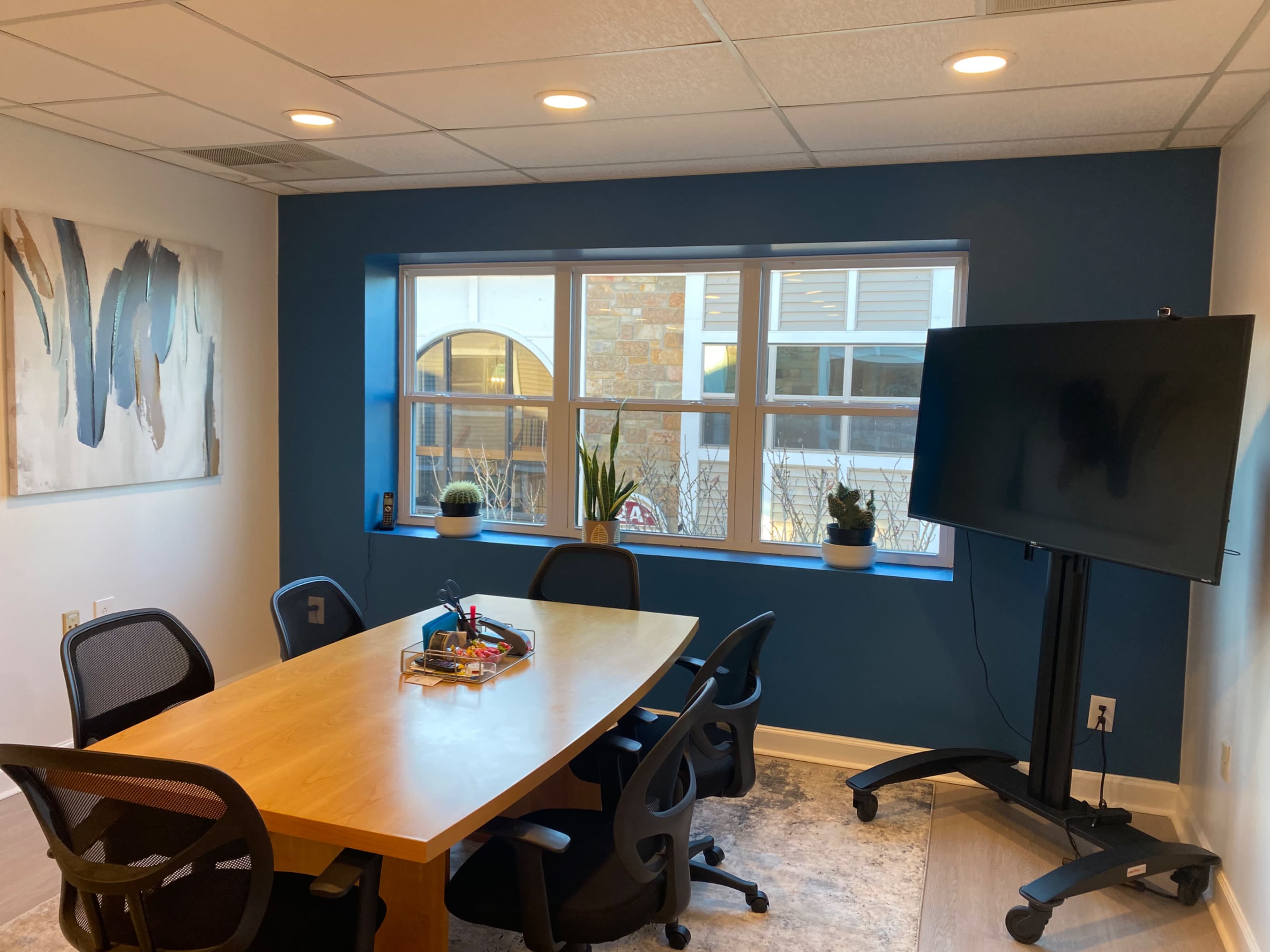 The image shows a modern conference room featuring a large wooden table surrounded by black office chairs, with a wall-mounted screen on a mobile stand and a window providing natural light.