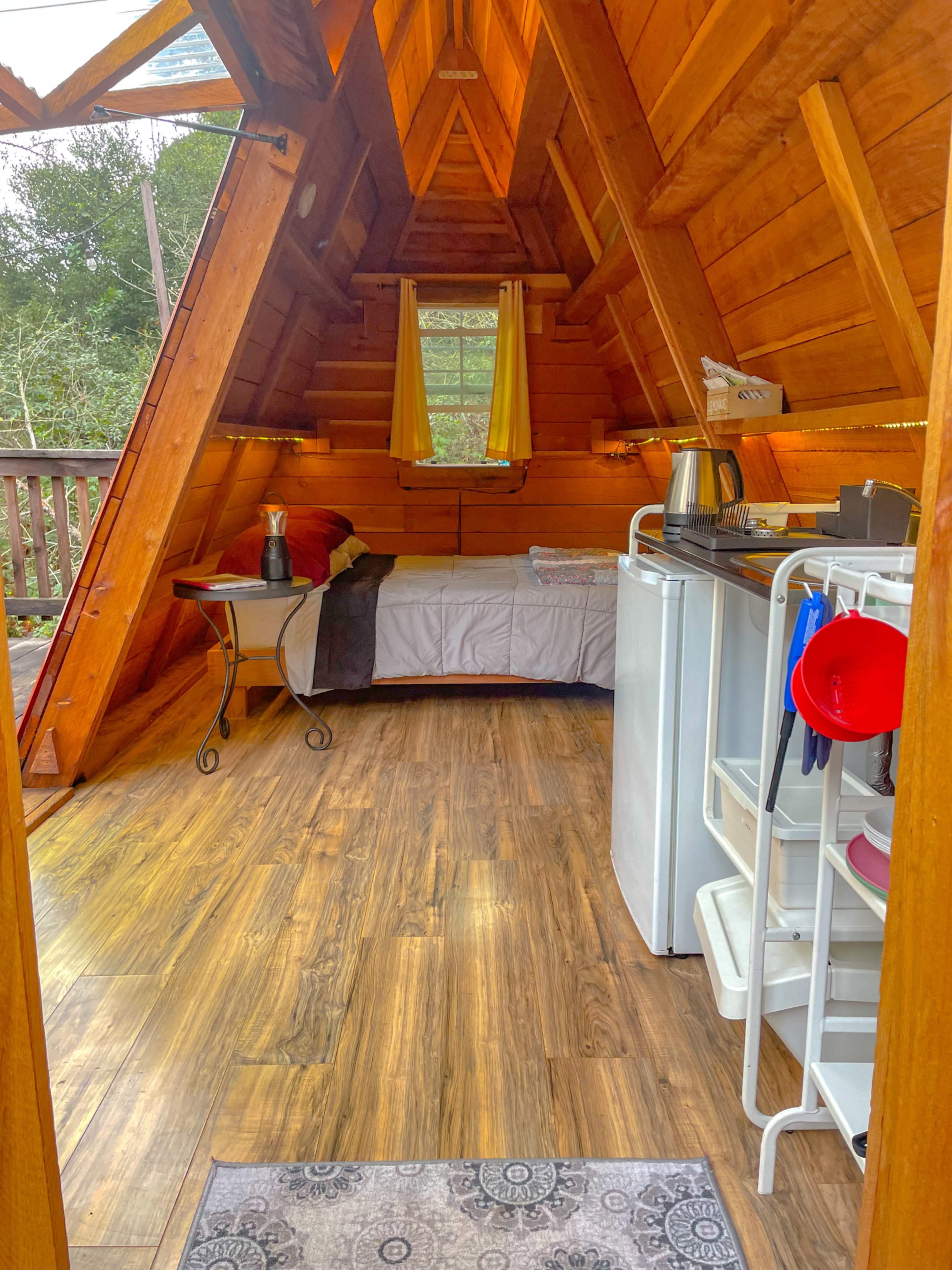 The interior of an A-frame cabin features wooden walls, a bed in the corner, a small kitchen area, and a table with a decorative vase.