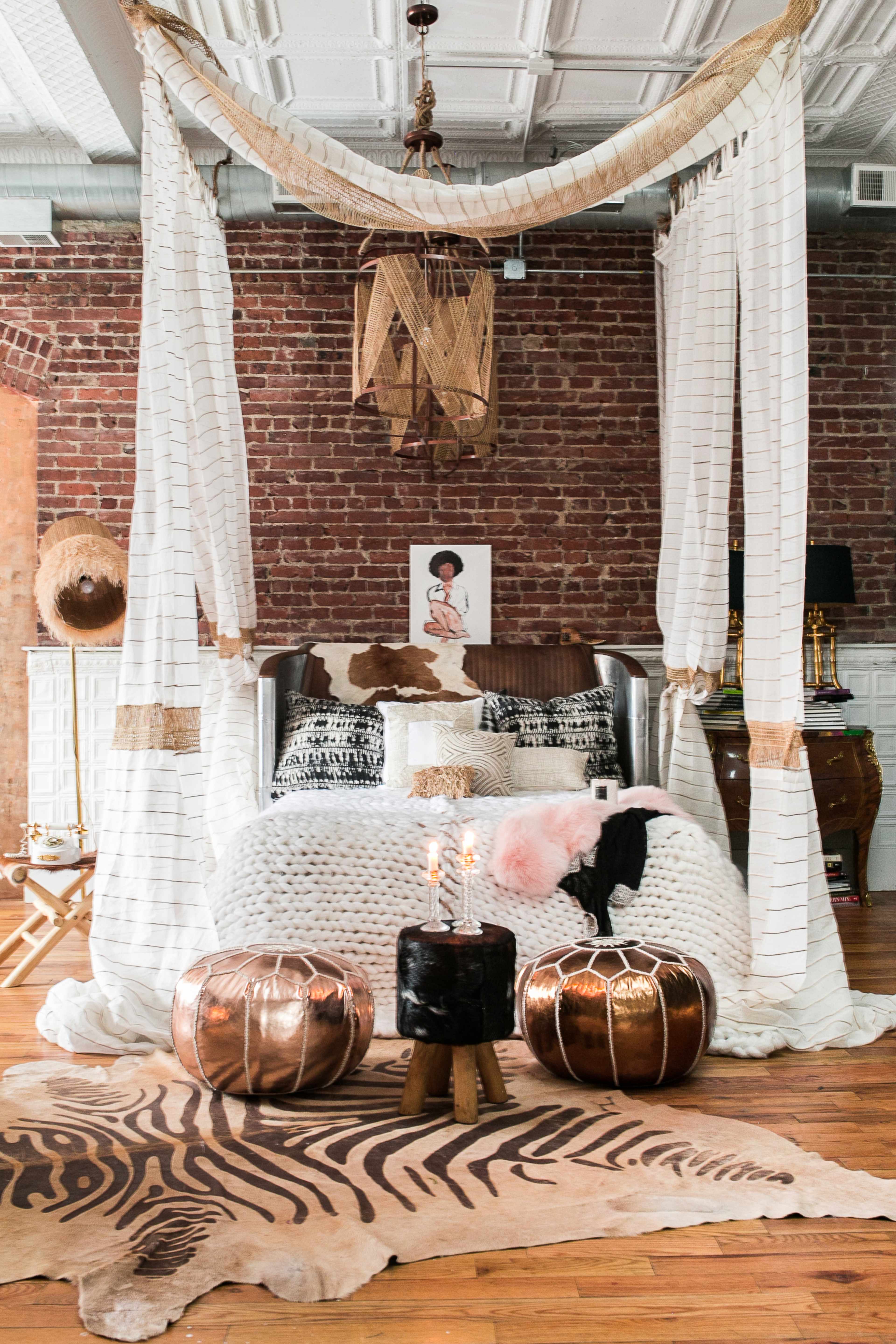 A cozy bedroom features a large bed draped with white fabric, surrounded by decorative pillows, a patterned rug, and two metallic poufs.