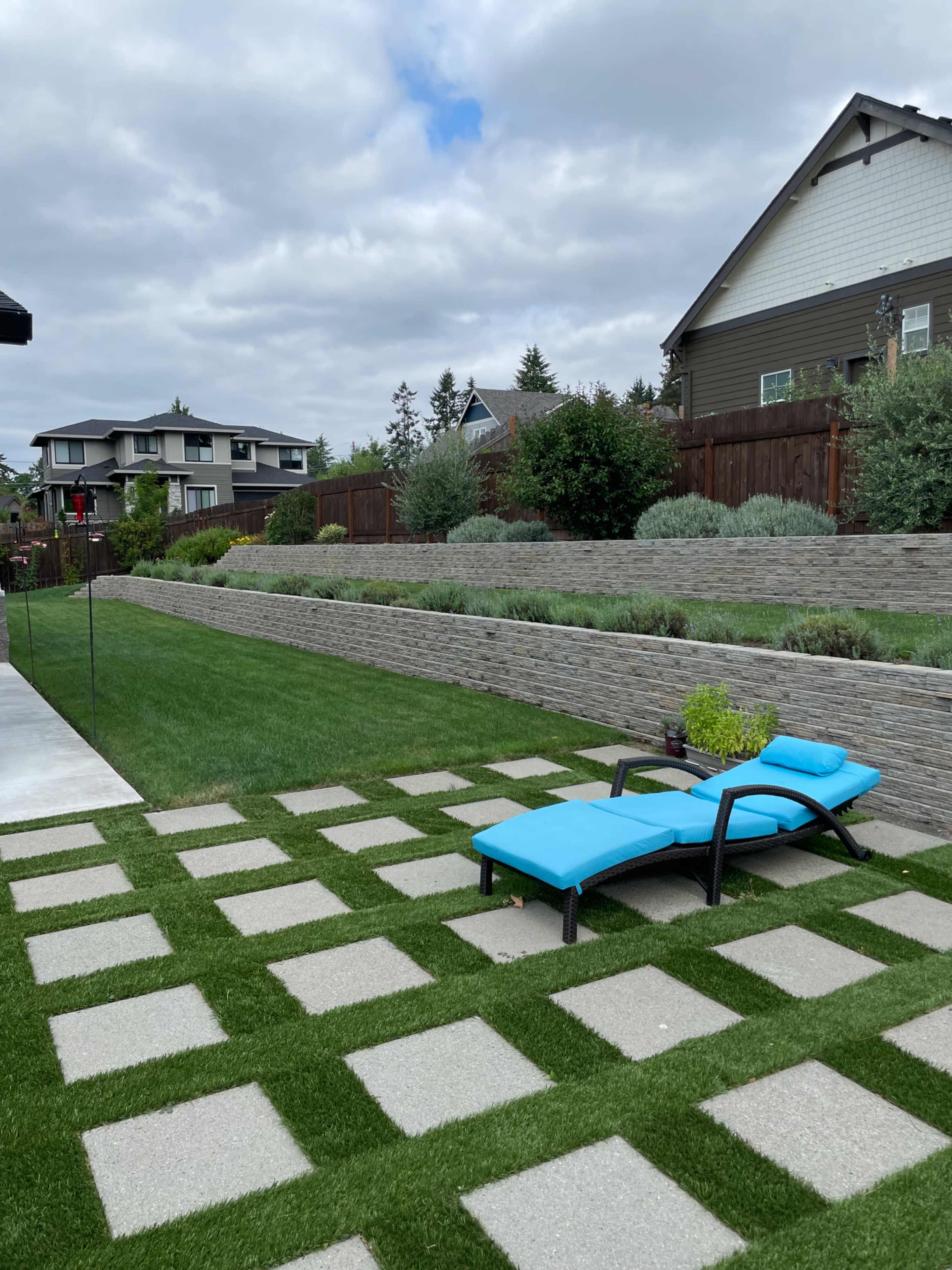 A landscaped backyard with a concrete paver pathway, a blue lounge chair, and neatly trimmed grass bordered by stone walls and shrubs.