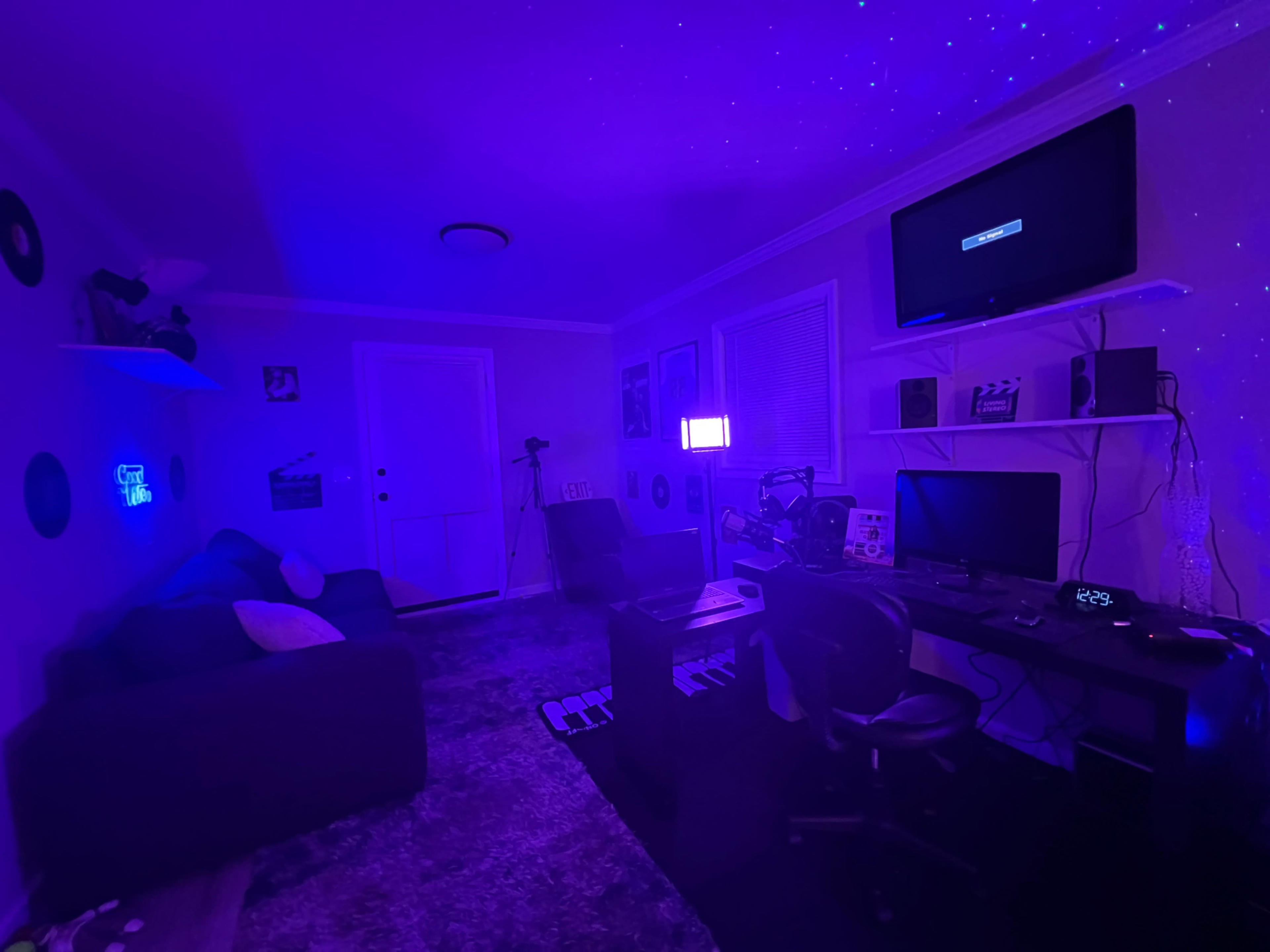 The room features a dark purple ambiance with a couch, desk setup, and a television displaying a black screen.