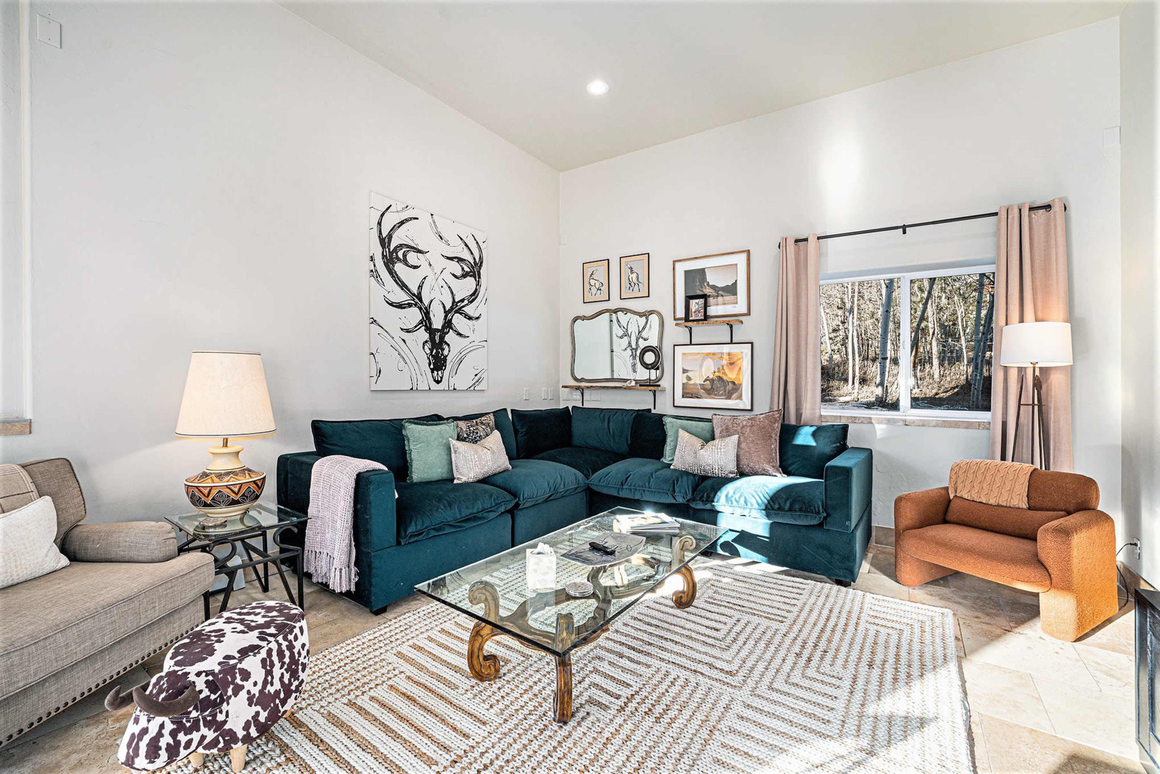 The image shows a modern living room featuring a large teal sofa, a glass coffee table, various framed artworks on the walls, and a mix of seating options including an orange chair and a beige armchair.