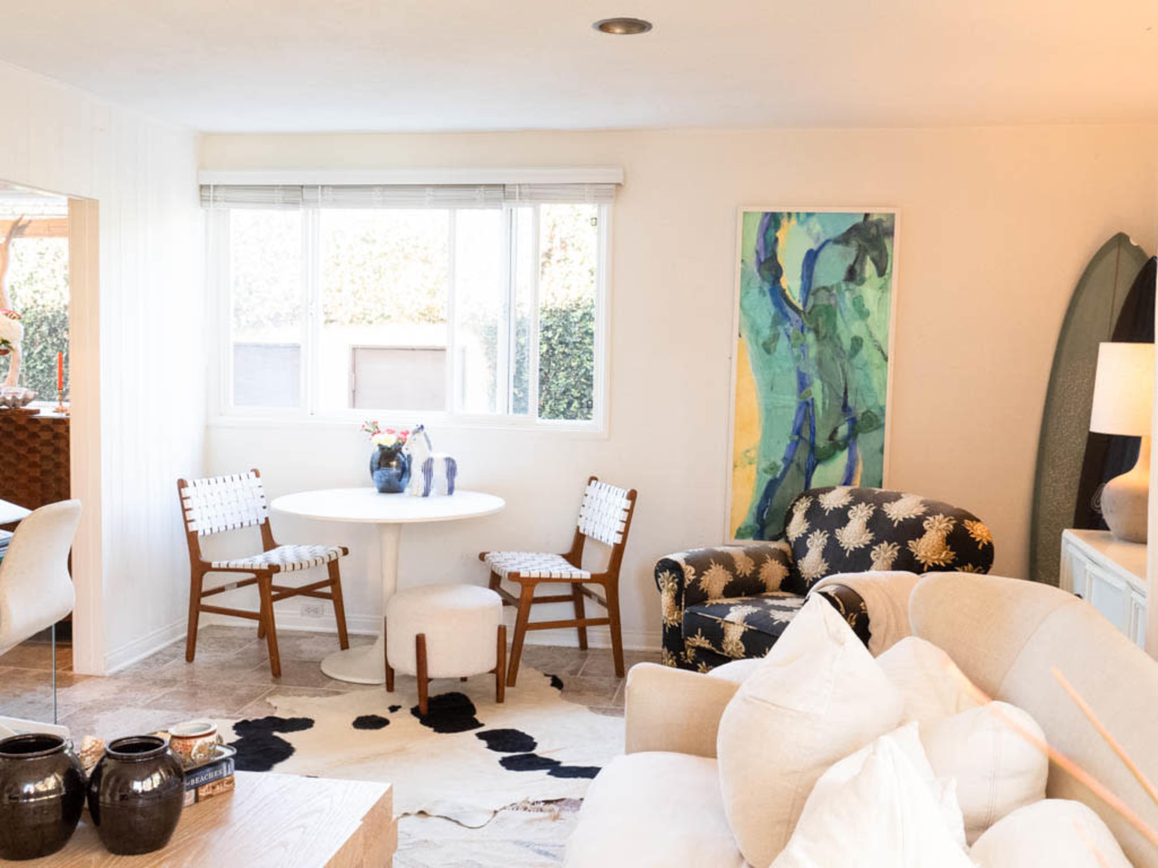 A bright living room features a round white table with two chairs, a patterned sofa, and an abstract painting on the wall.