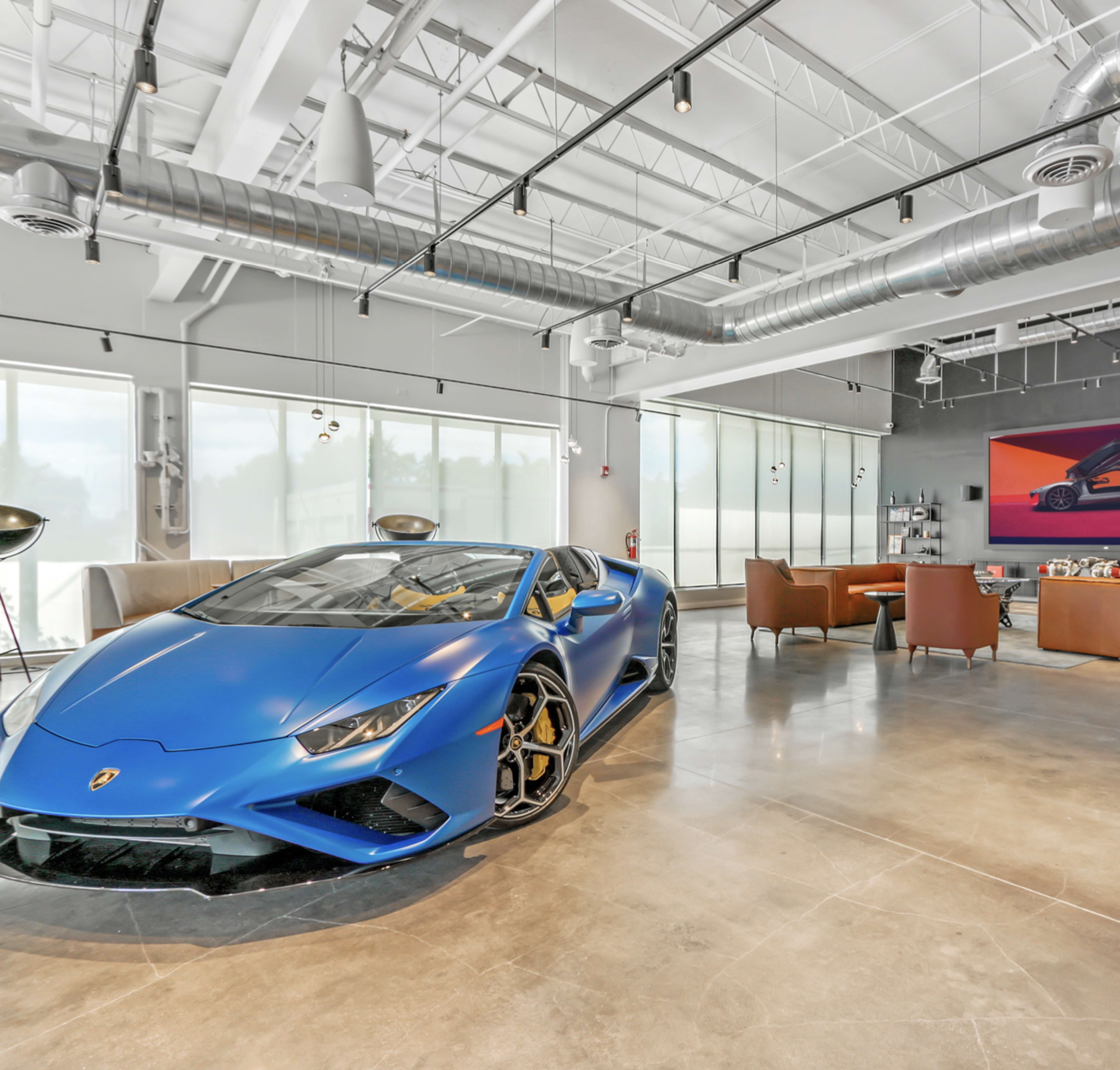 A blue Lamborghini is prominently displayed in a modern showroom with large windows and contemporary seating.