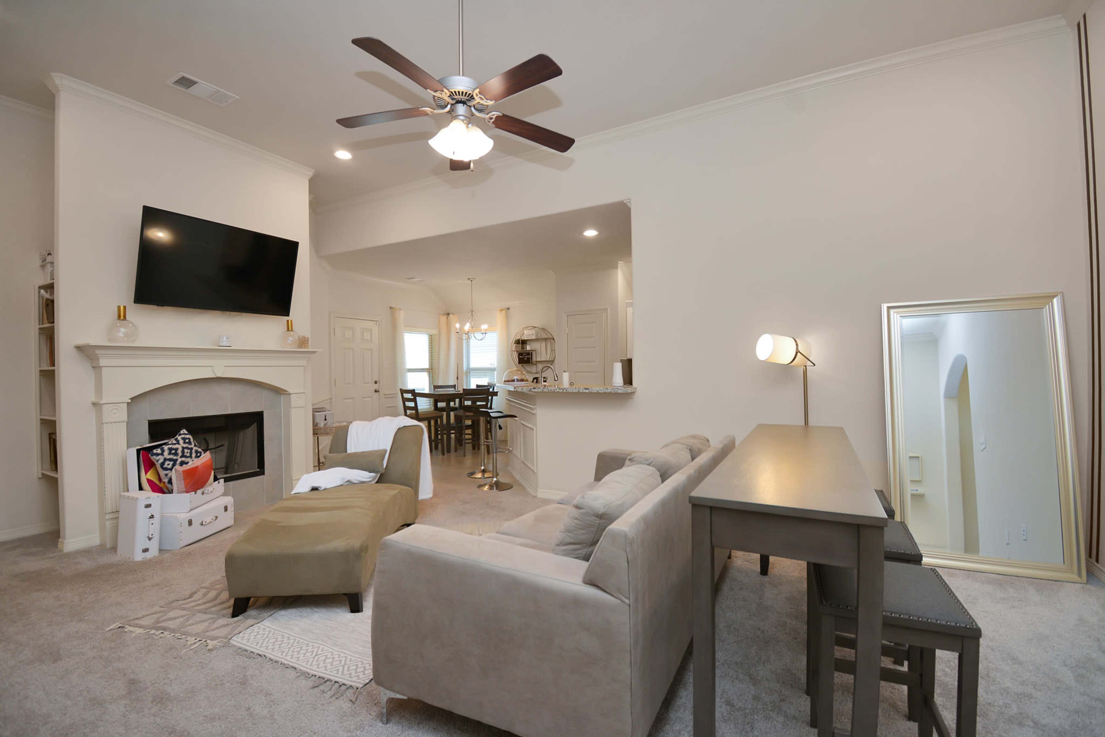 A living room with a beige sofa, a mounted television above a fireplace, and a dining area visible in the background.