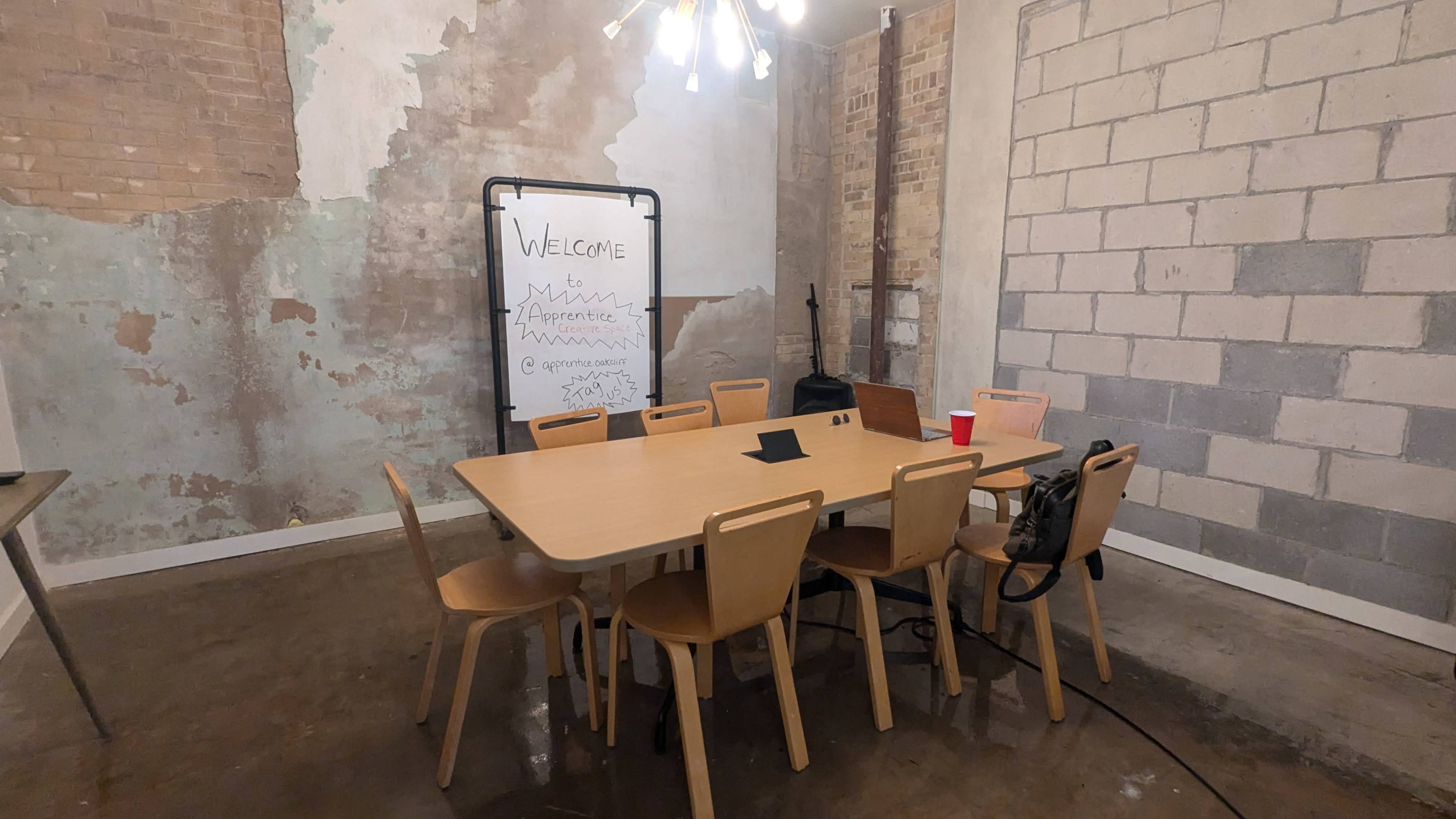 A meeting room features a large table surrounded by chairs, with a chalkboard displaying a welcome message on one wall and exposed brick and concrete surfaces.