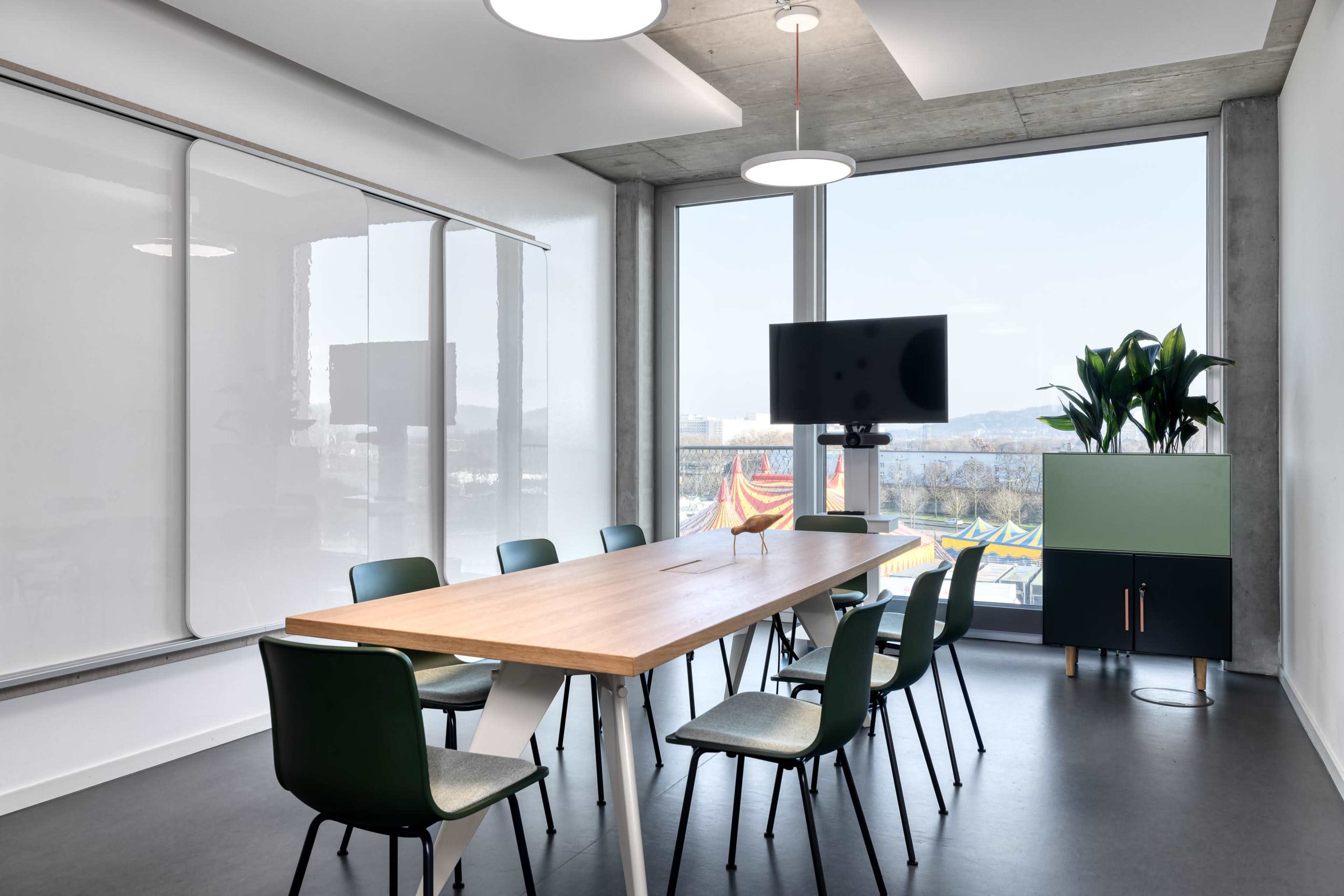 A modern conference room with a long wooden table surrounded by green chairs, a television on the wall, and large windows overlooking an outdoor area.