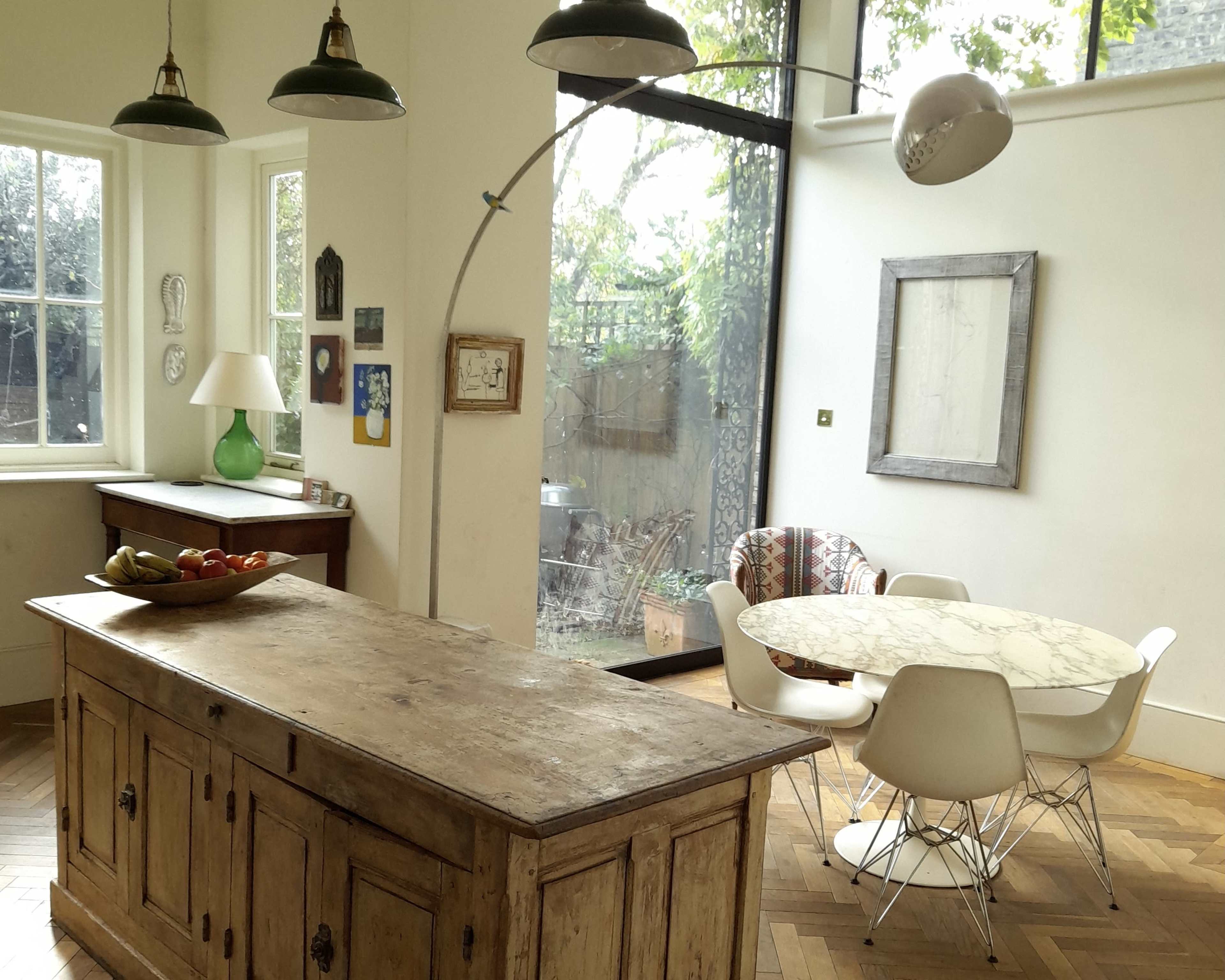 A kitchen features a wooden island with a fruit bowl, a round marble table with white chairs, and large windows that provide a view of the garden outside.