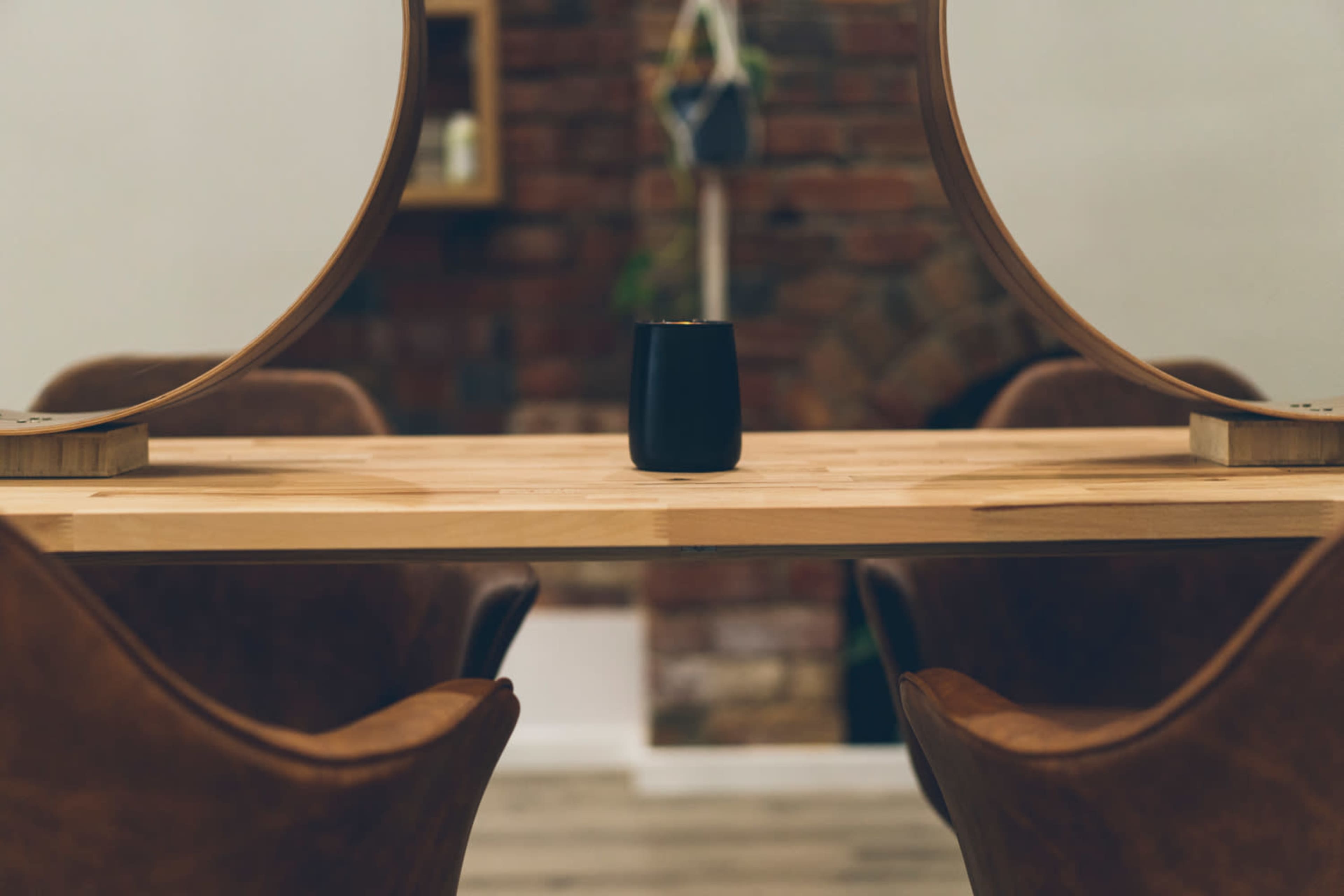 A wooden table with a black candle sits between two curved mirrors in a brick-walled room.