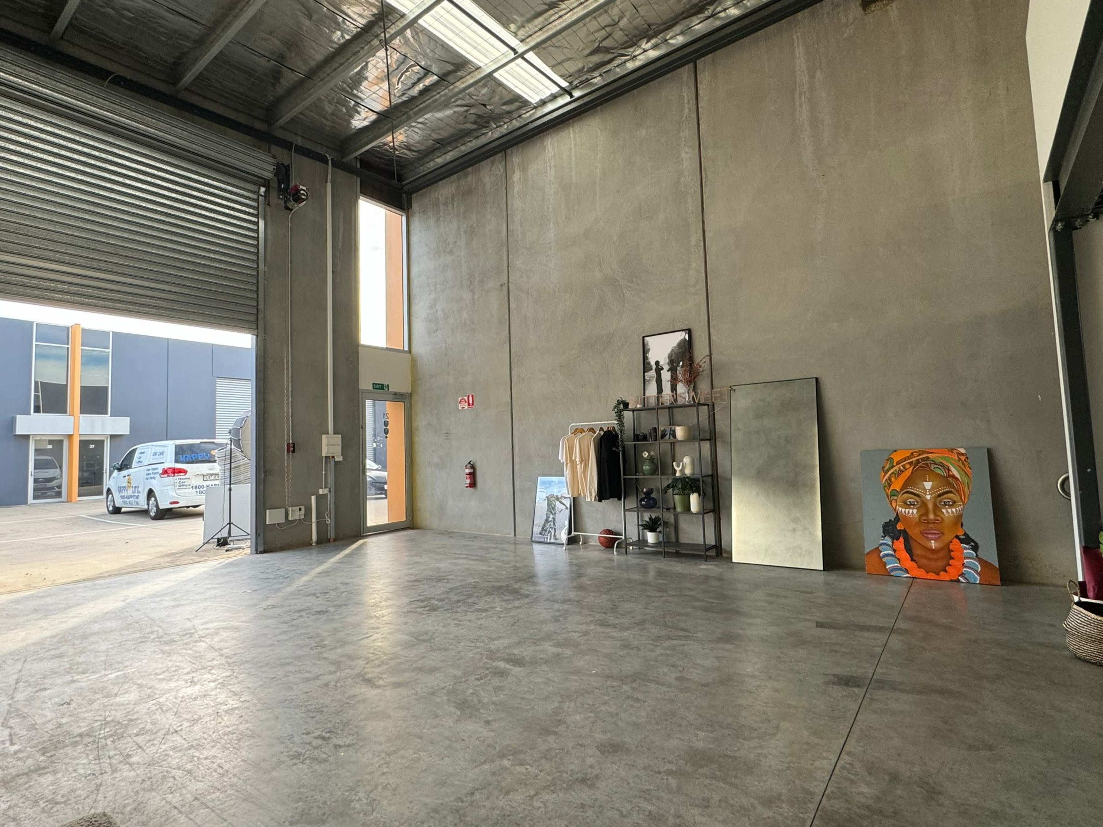 A spacious, bare concrete interior of a building with a large roll-up door, a display shelf holding clothes and decor, a standing mirror, and a colorful artwork of a woman on the floor.
