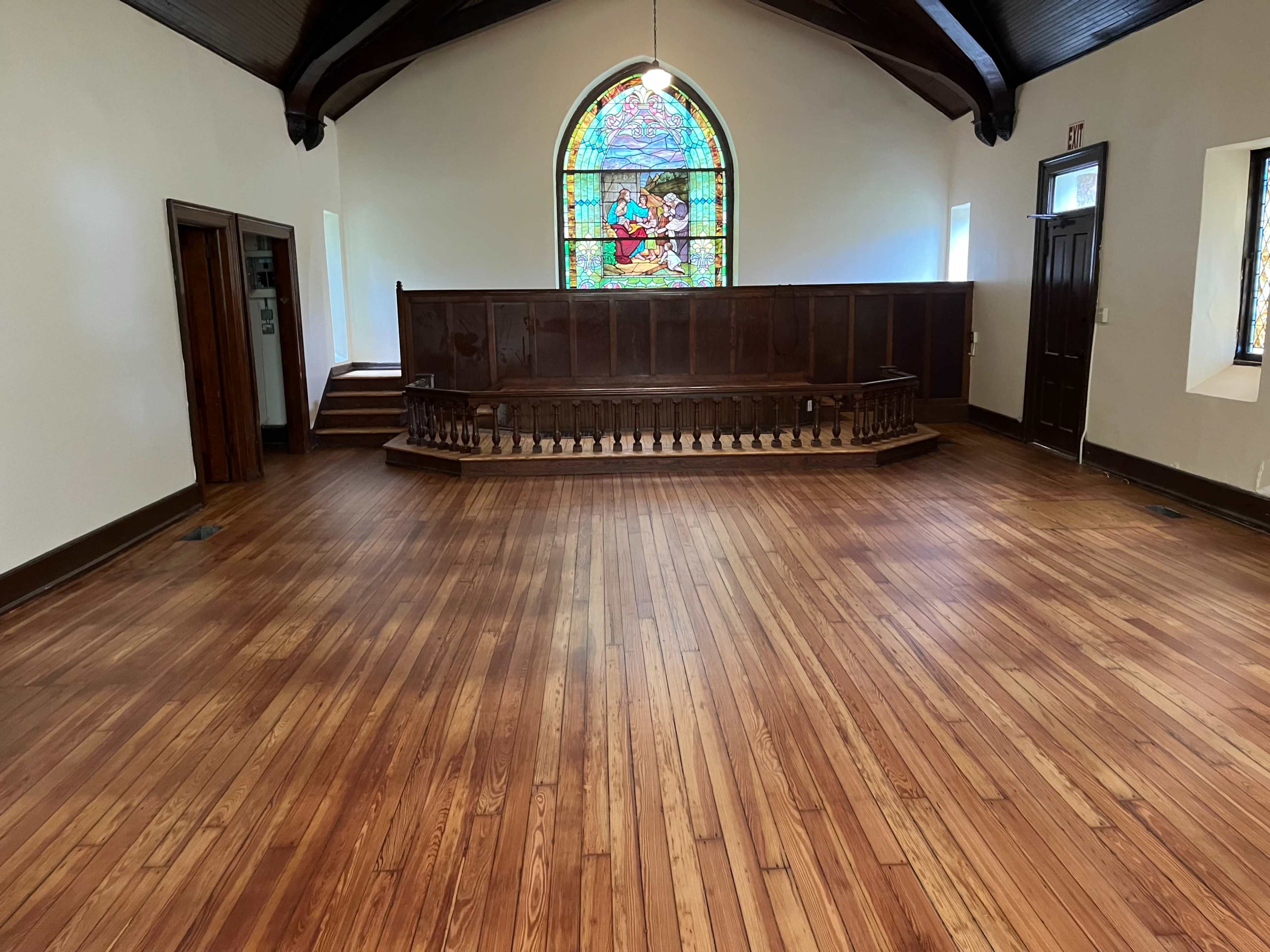 The image shows an empty room with wooden flooring, a raised platform at the back, and a colorful stained glass window.